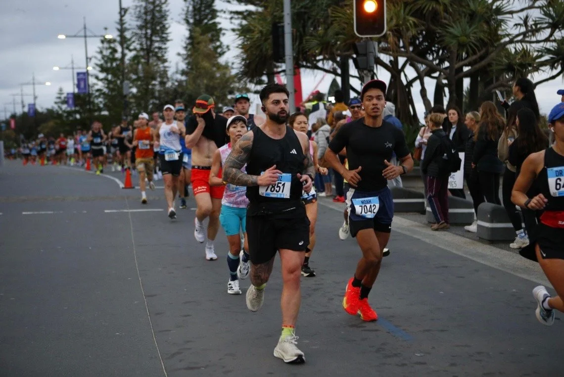 Group of runners participating in a marathon on a city street with spectators along the sidewalk and trees in the background.