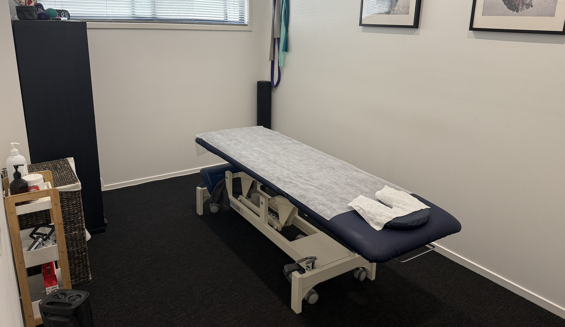 Empty medical examination or therapy room with a treatment table covered in white paper, a small rolling cart with supplies, and framed pictures on the wall.