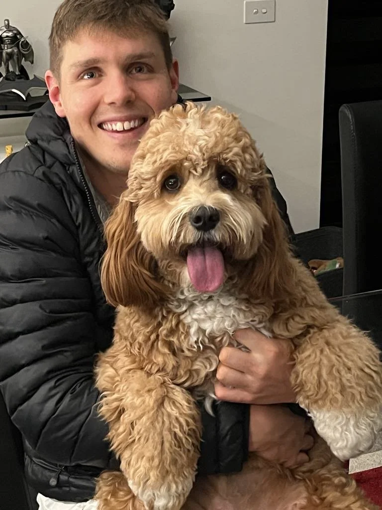 A man smiling and holding a fluffy, tan and white poodle mix dog with its tongue out
