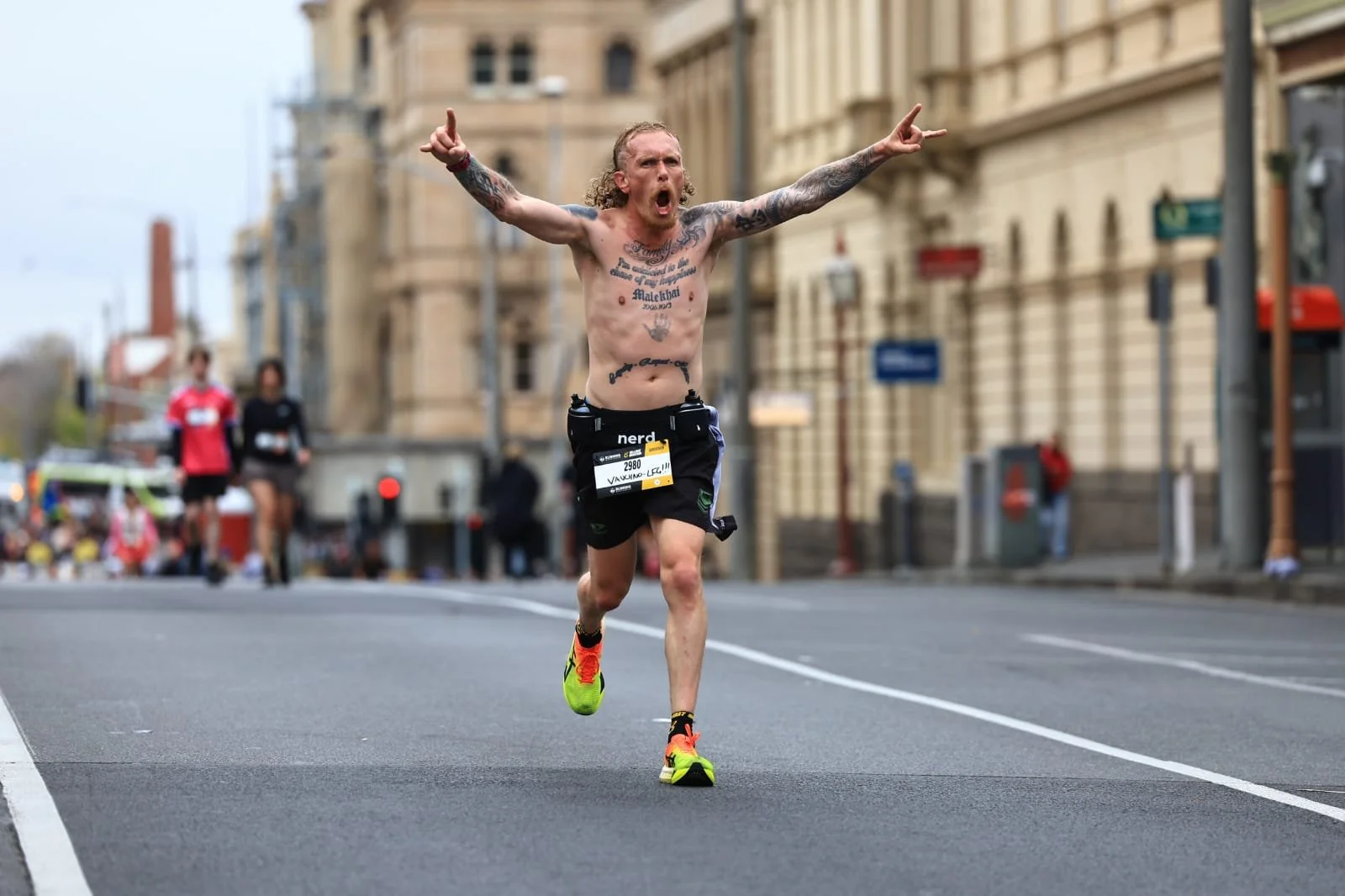 Tattoed man running shirtless in a marathon with his arms raised, middle of street in an urban area, other runners following in the background.