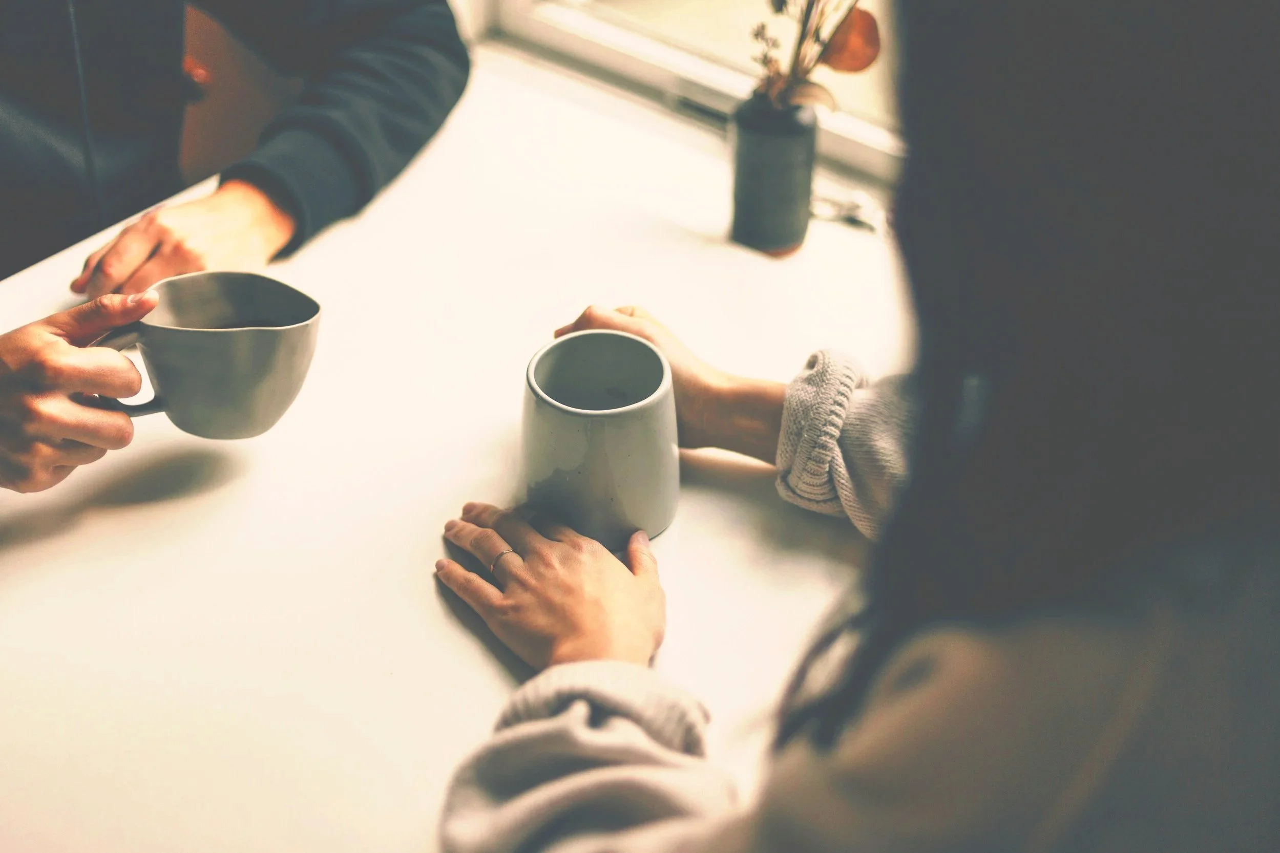 Two people exchanging coffee mugs at a table with natural light coming through a window.
