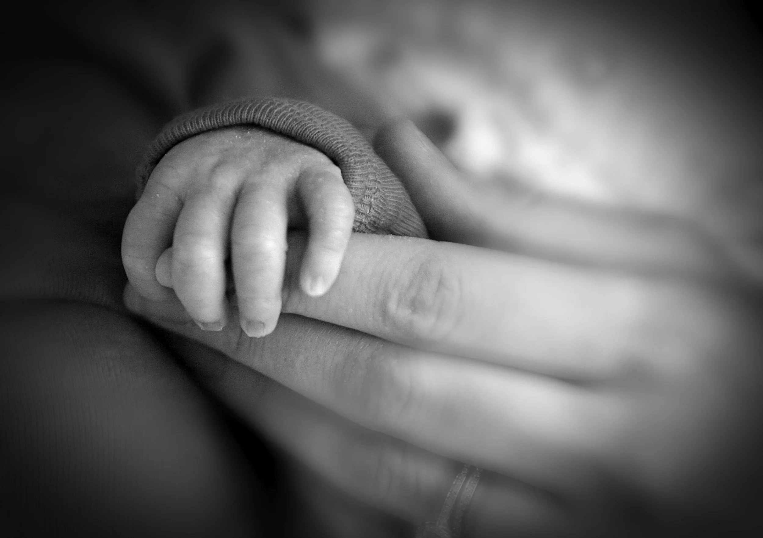 Black and white photograph of a hand gently holding a child's hand, focusing on the hands with a blurred background.