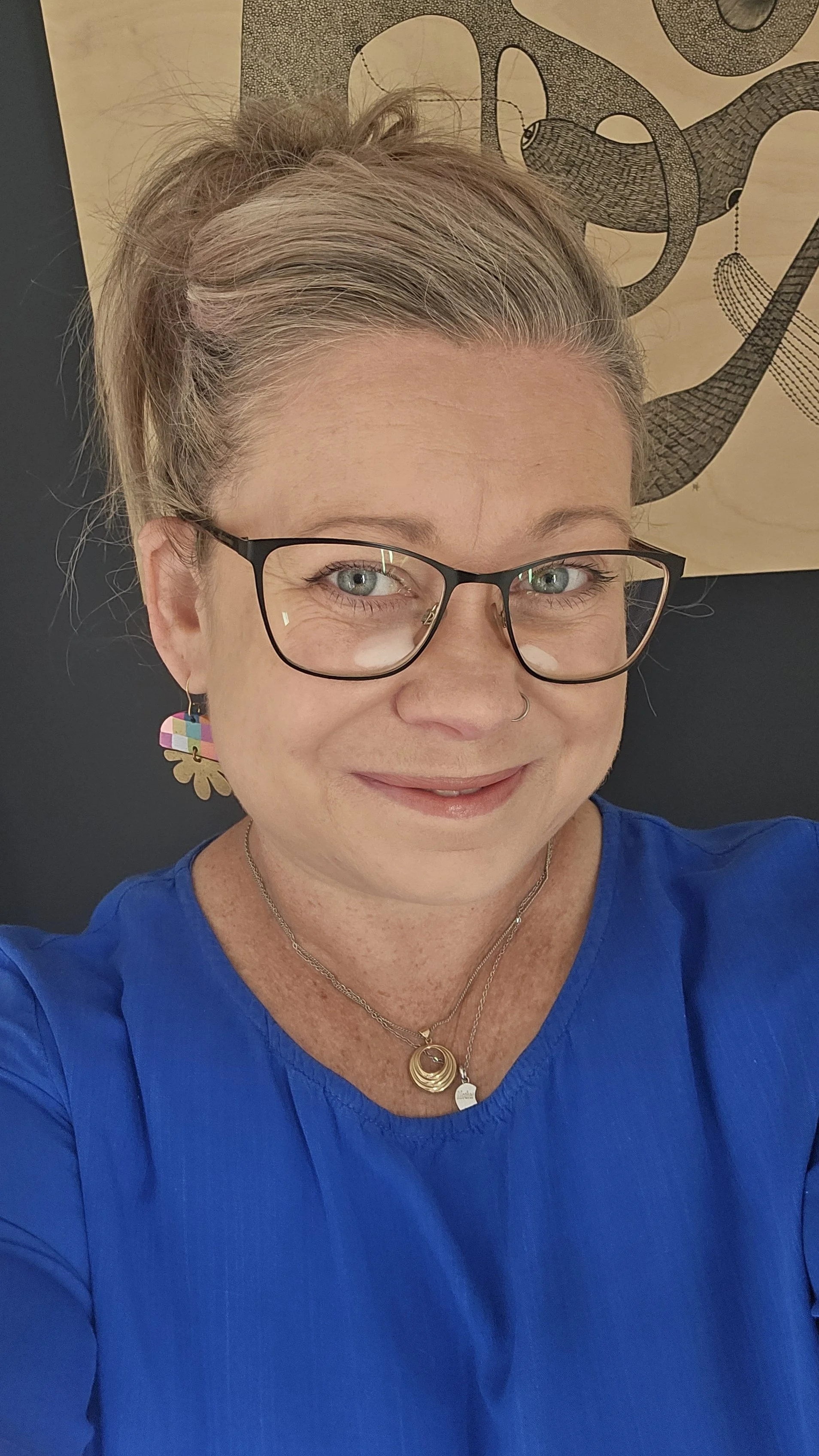 A woman with short hair, wearing glasses and colorful earrings, smiling in a blue shirt, with a piece of artwork featuring a snake in the background.