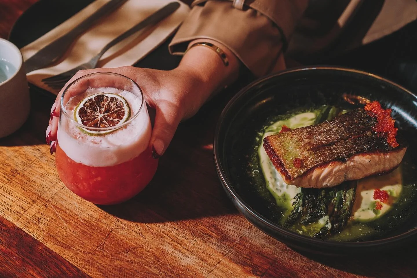 A person's hand holding a glass of pink cocktail with a slice of dried citrus fruit on top, next to a bowl of seared fish on a bed of sauce with green herbs and red roe, on a wooden table.