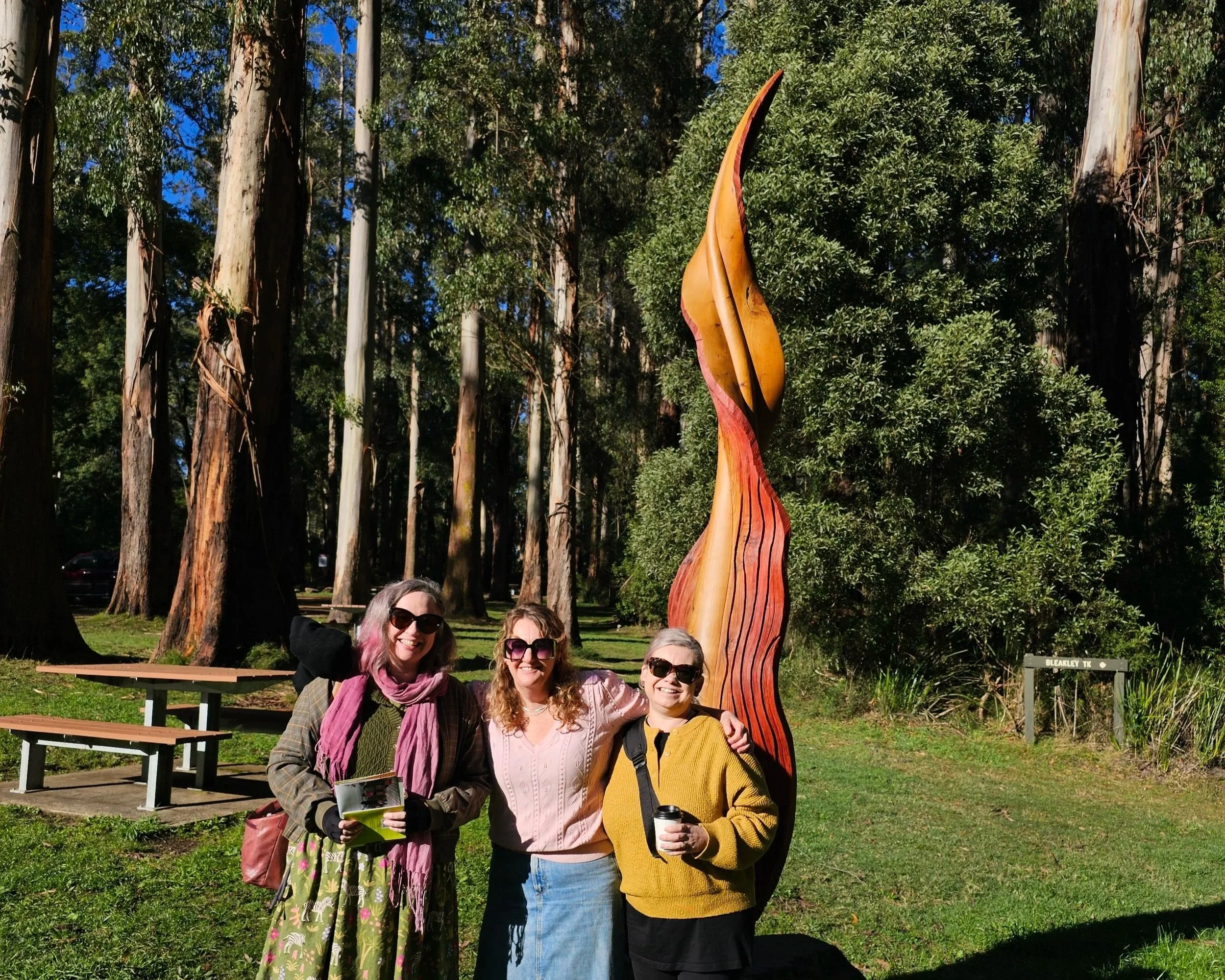 A group of five women posing together outdoors in front of a large wooden sculpture of a colorful, stylized bird or dinosaur on a sunny day with a woodland background.