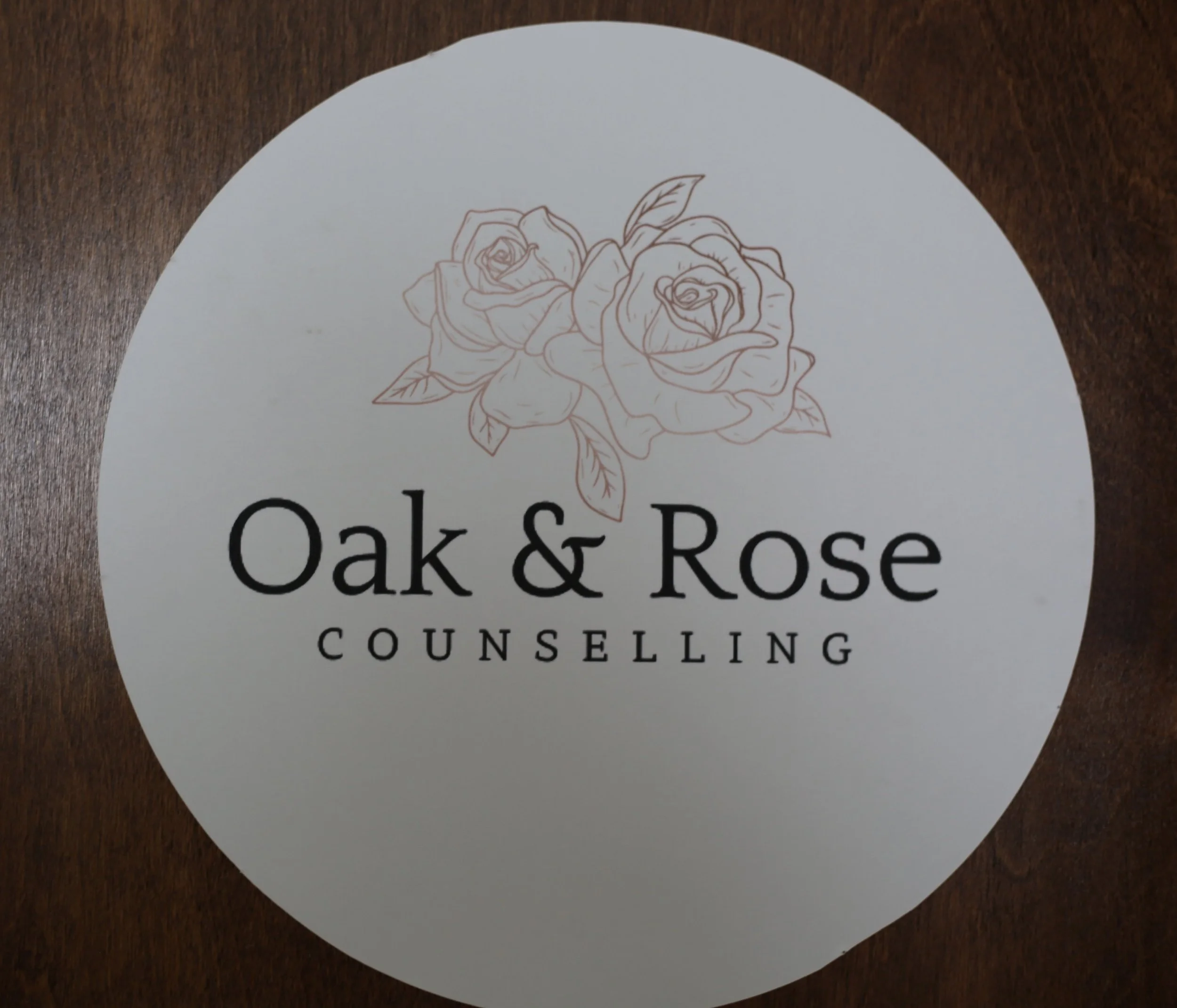 A round white sticker with a line drawing of roses and the words "Oak & Rose Counseling" on a wooden surface.