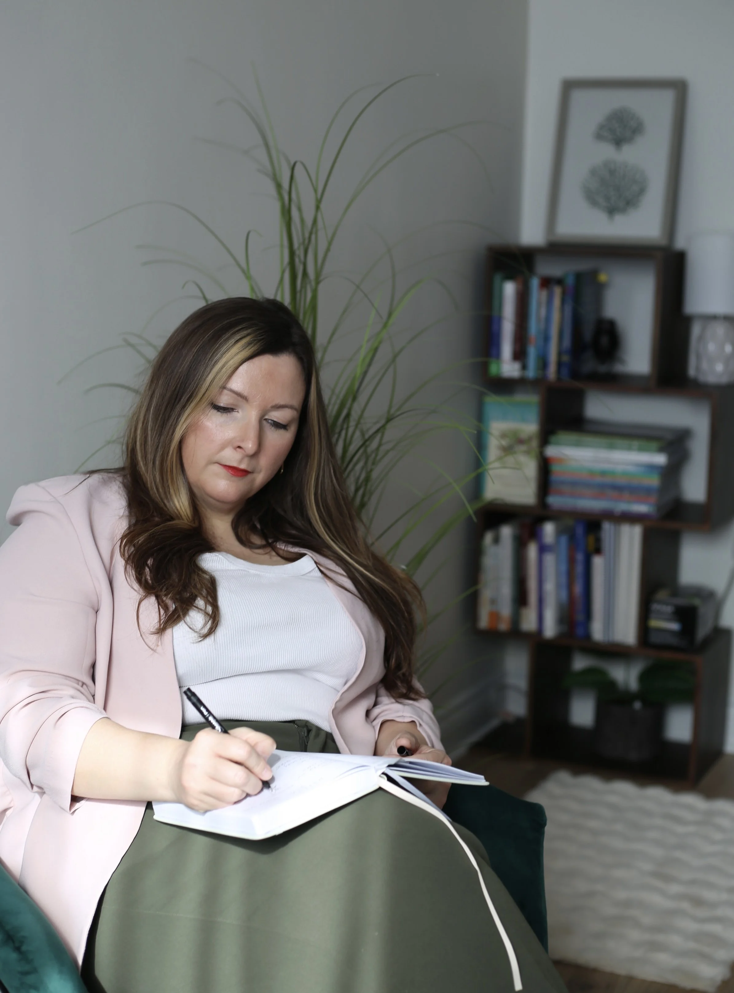 A woman with long hair, wearing a white top and pink blazer, is sitting on a gray chair, writing in a notebook with a black pen in a cozy room with a bookshelf and framed art of two trees in the background.