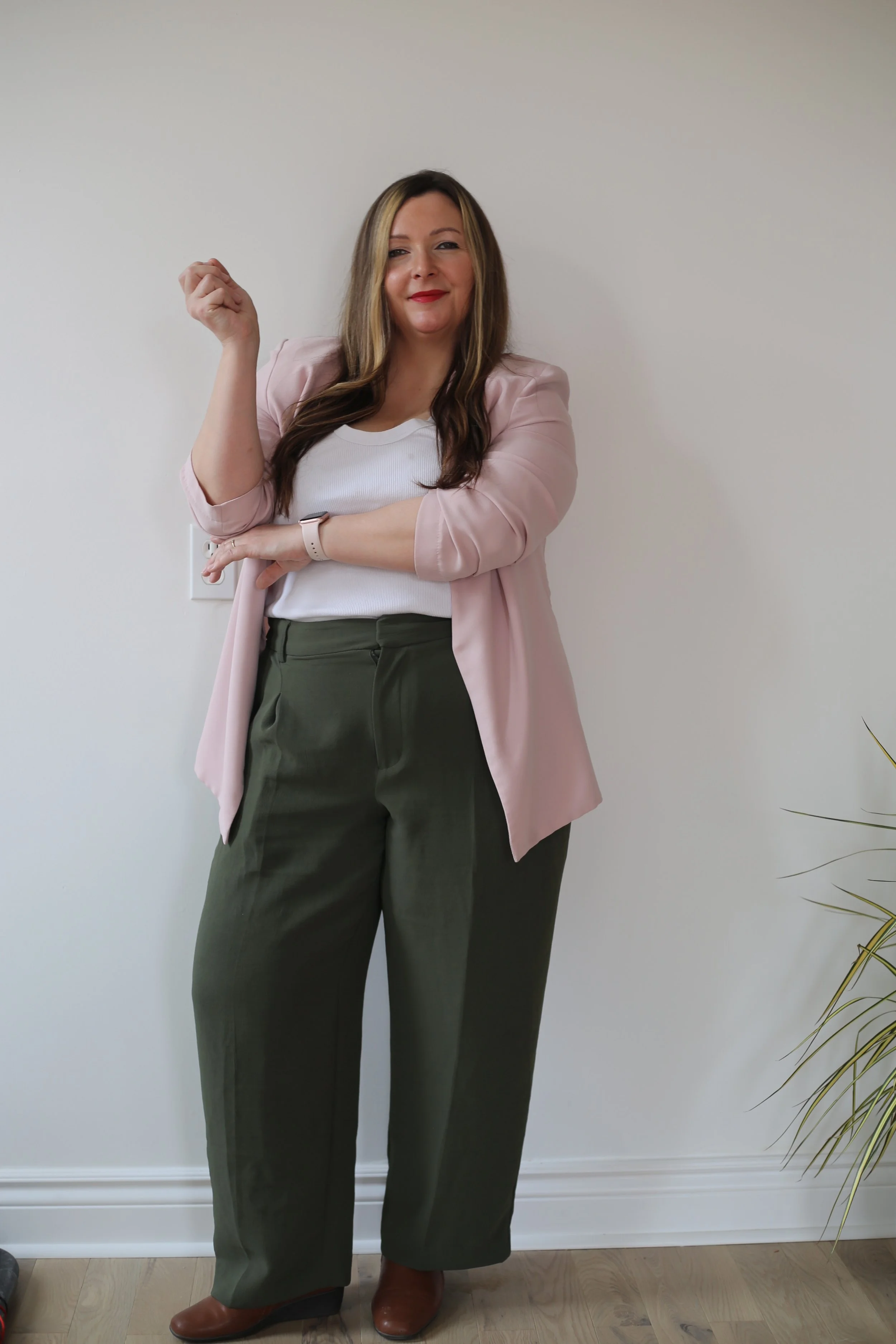 Woman with long brown and blonde hair, wearing a pink blazer, white shirt, dark green pants, and brown boots, standing against a plain white wall and a plant are visible on the side.