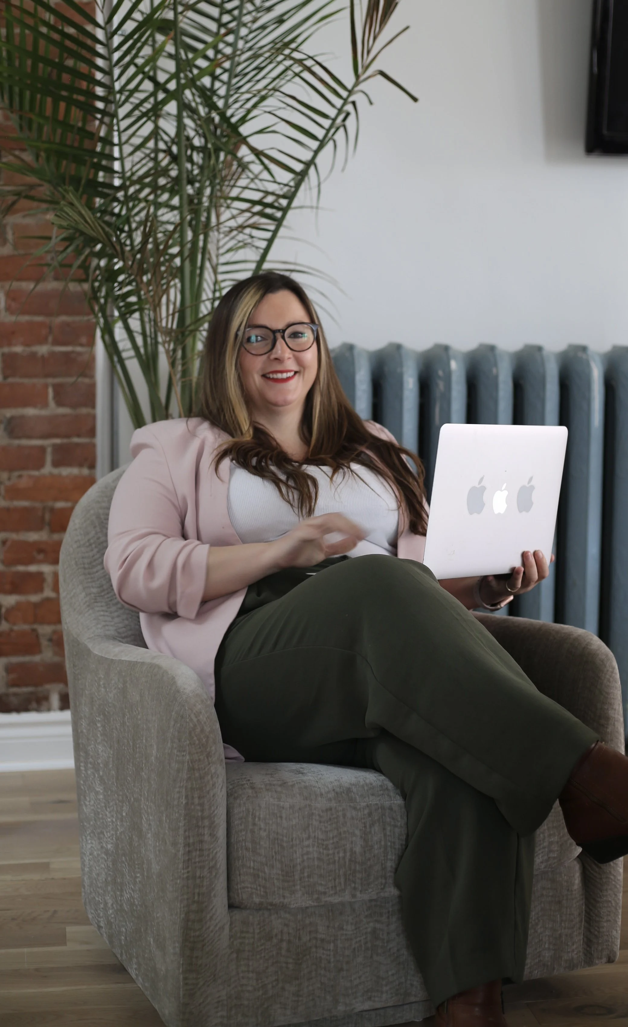 A woman with glasses, wearing a light pink blazer and white top, sitting on a beige armchair, holding a silver laptop with an Apple logo, smiling in a room with a brick wall, large green plant, and white wall with a radiator.