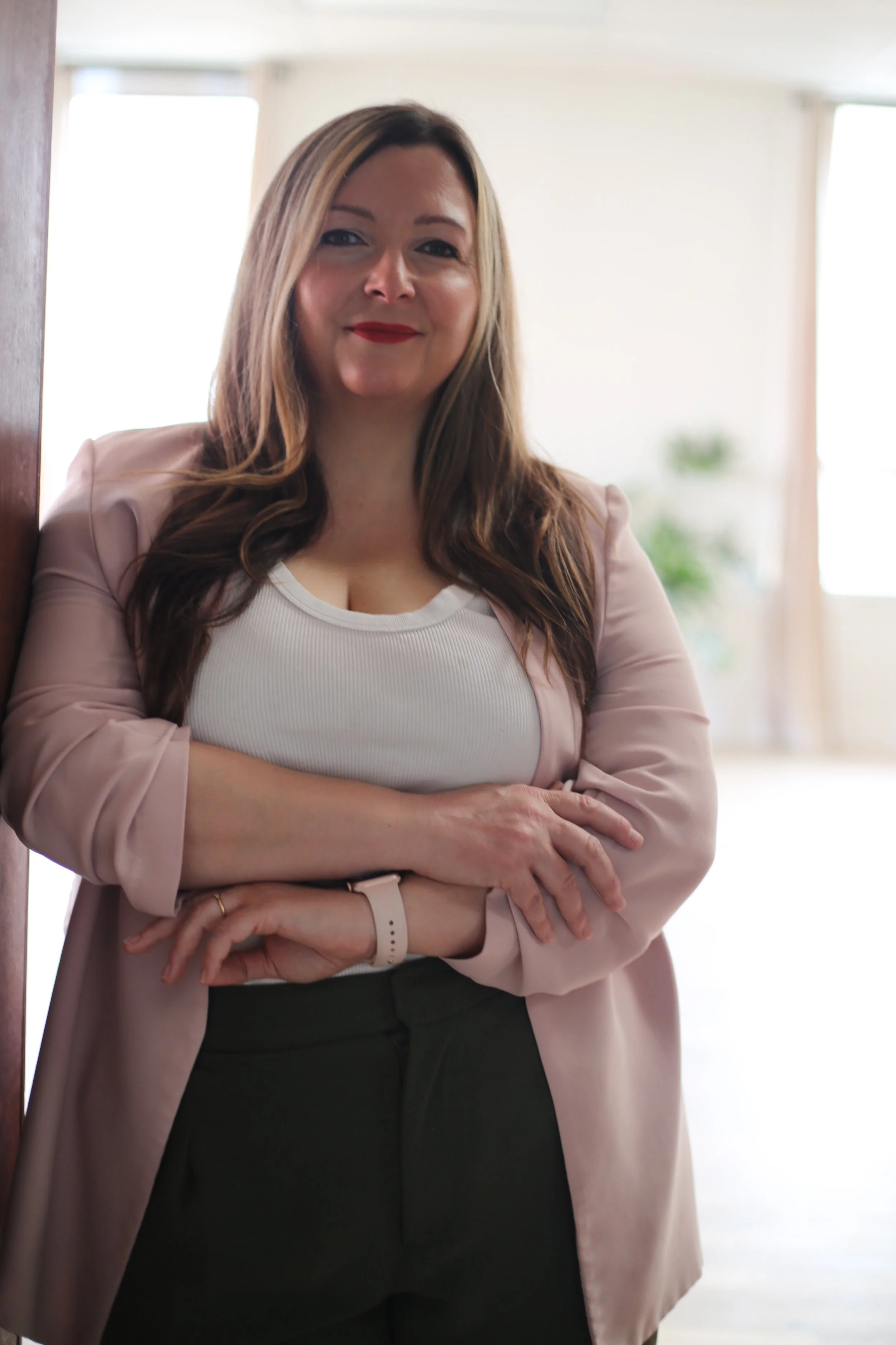 A woman with long, wavy, brown hair and red lipstick standing indoors with her arms crossed, wearing a white top, pink blazer, and black pants, in front of a light-colored wall and windows.