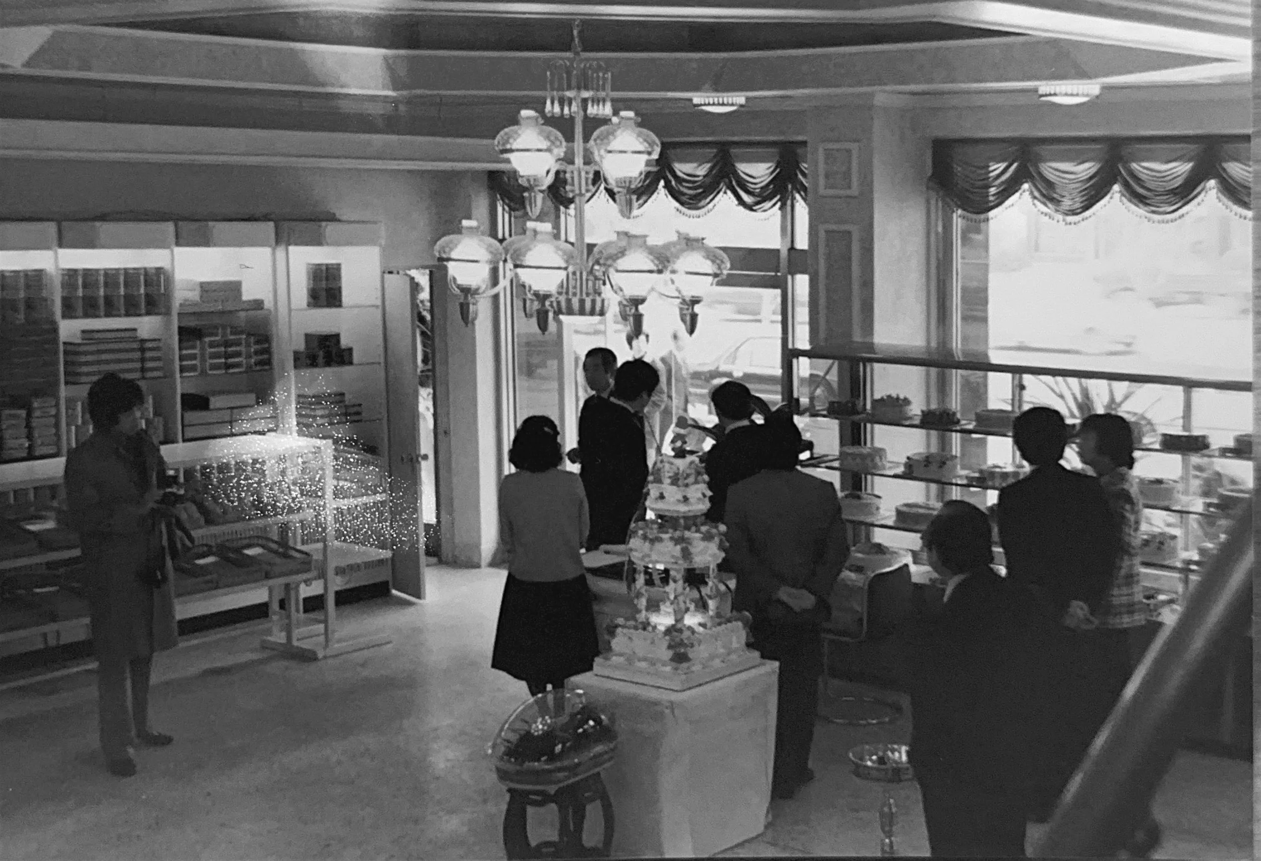 초창기 로얄제과 베이커리 내부에서 구경하는 사람들, 진열된 베이커리 선반과 큰 창, 샹들리에가 보이는 장면