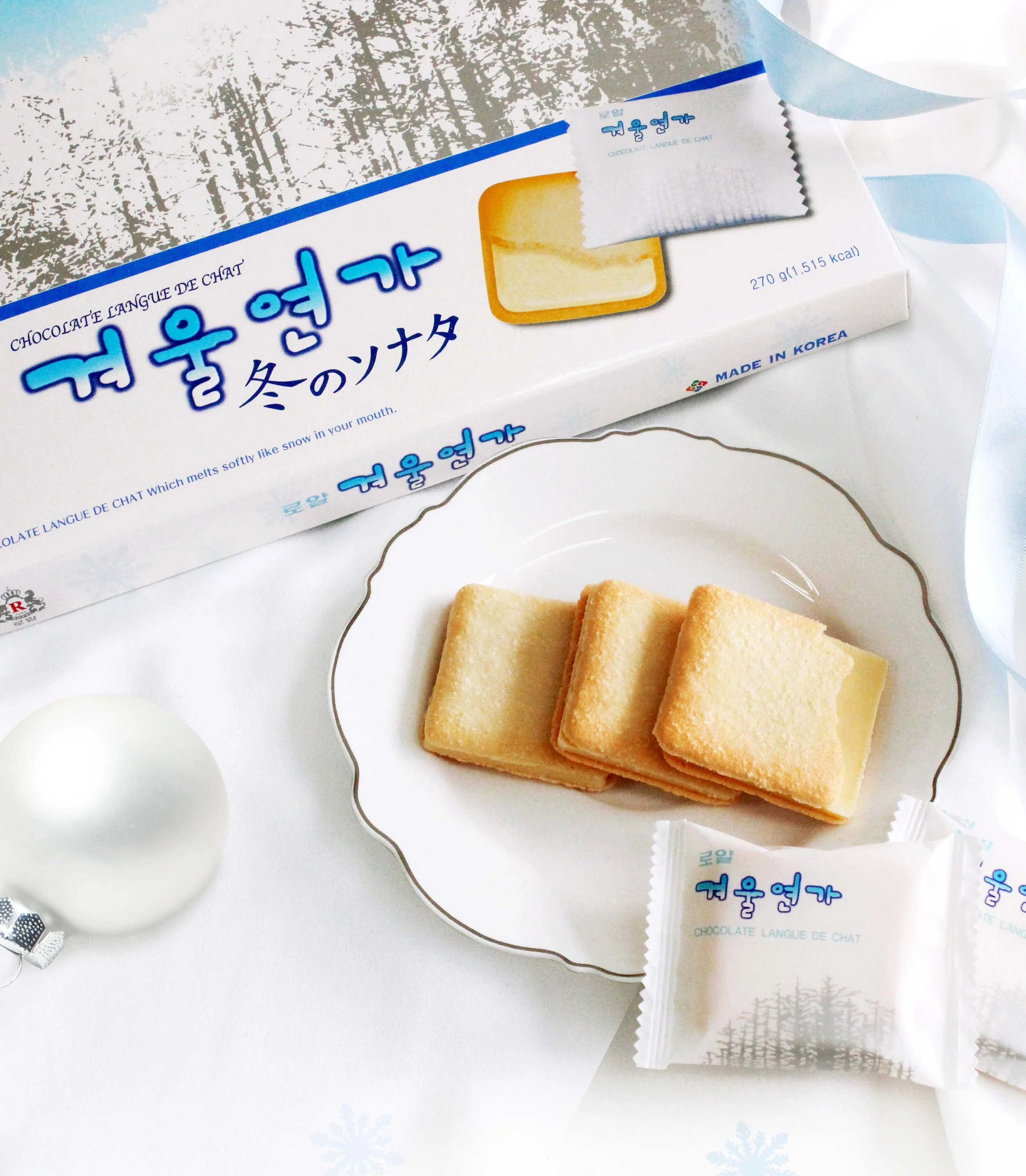 A box of Korean chocolate wafers labeled 'Chocolate Language de Chat' with Korean text, a slice of the wafer on a decorated white plate, a wrapped wafer snack, and a white Christmas ornament, all on a white table with winter-themed snowflake decorations.