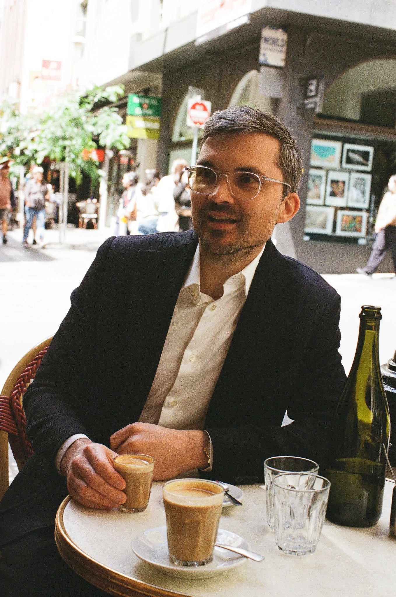 A man with glasses in a black suit jacket and white shirt sitting at a table outside, holding a glass of coffee, with two other coffee glasses, a large bottle, and empty glasses on the table, at an outdoor cafe with pedestrians in the background.