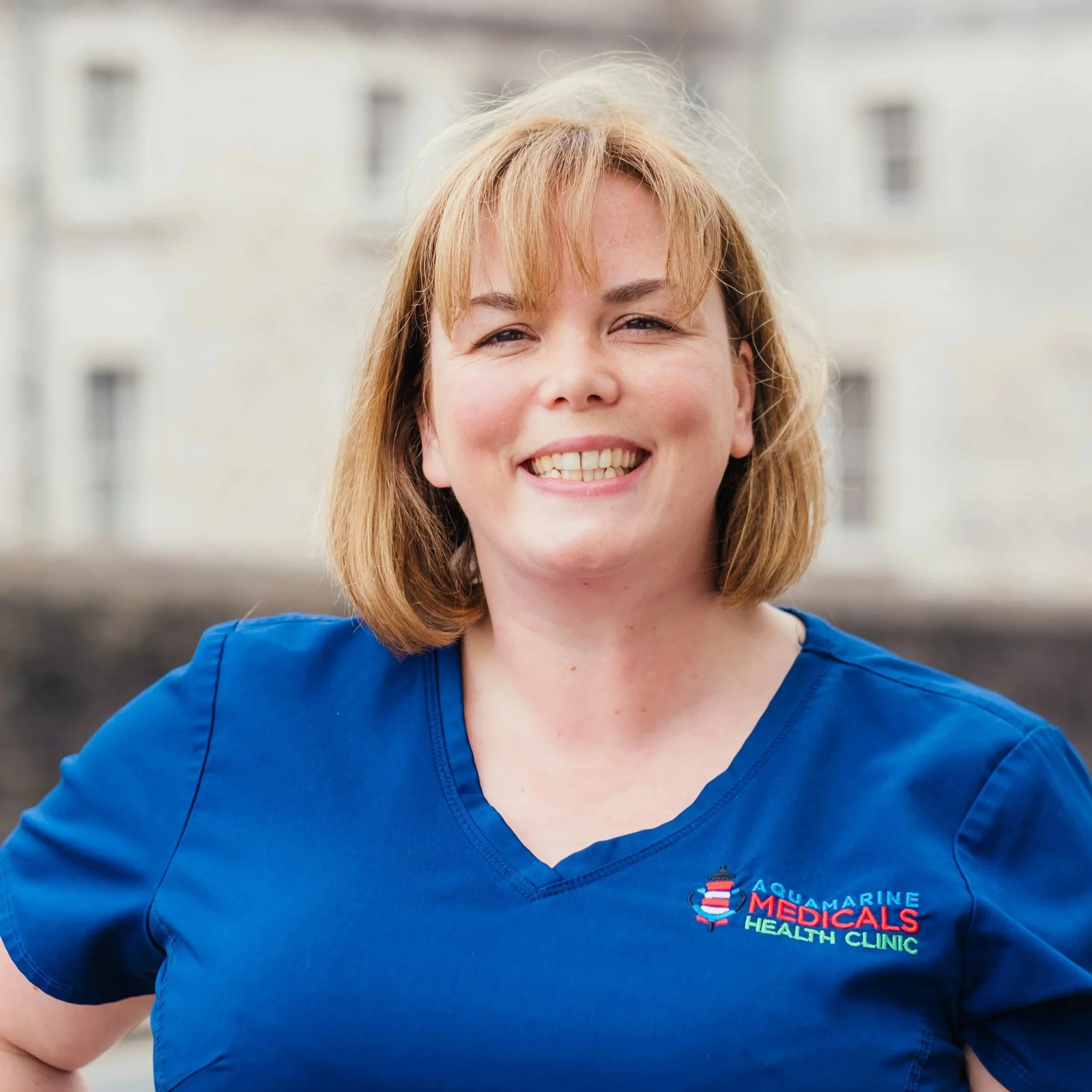 A woman with shoulder-length blonde hair wearing a blue Medicals Health Clinic uniform, smiling outdoors in front of a pale building.
