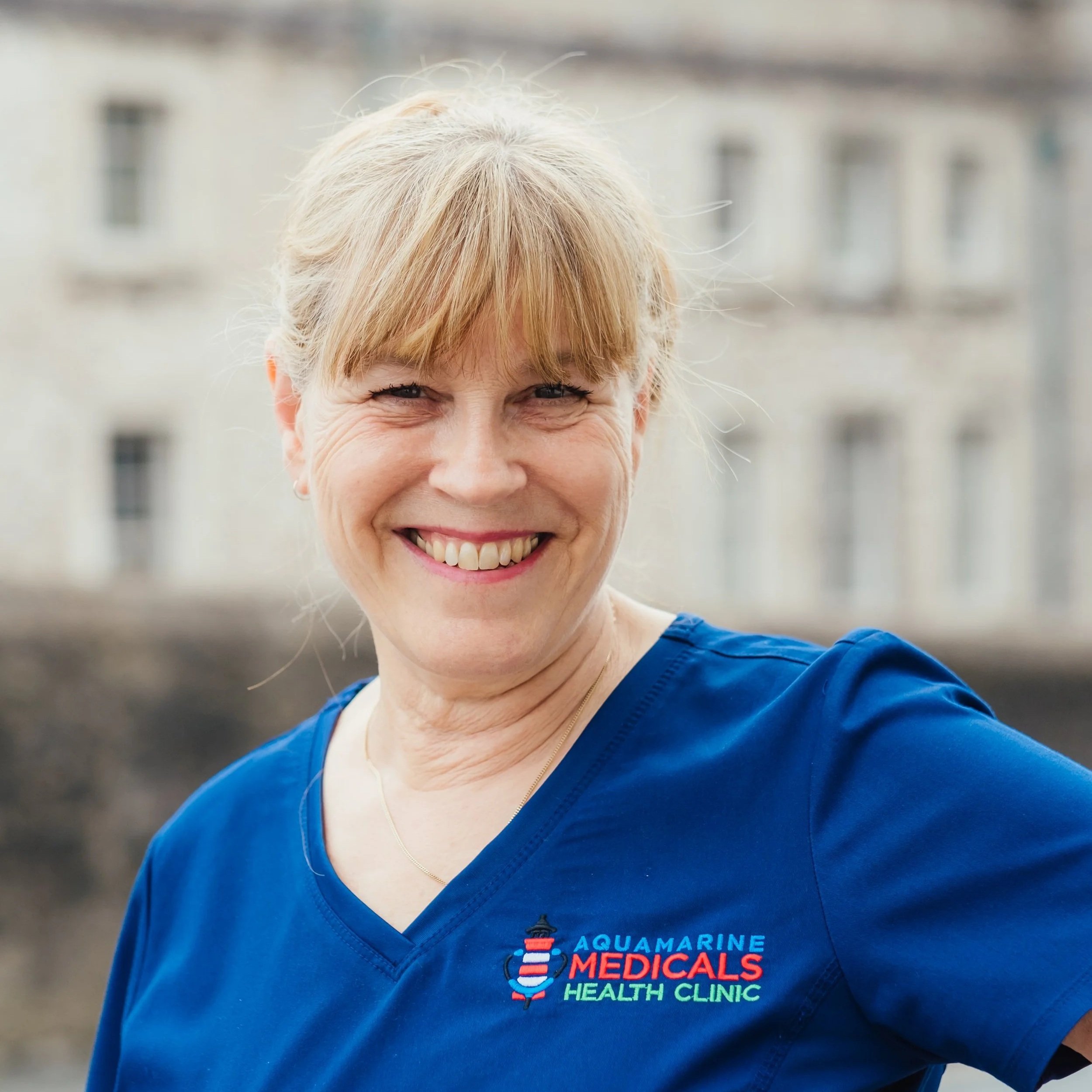 A smiling woman with blonde hair wearing a blue medical scrub top with the logo of Aquamarine Medicals Health Clinic. She is outdoors with a blurred building in the background.