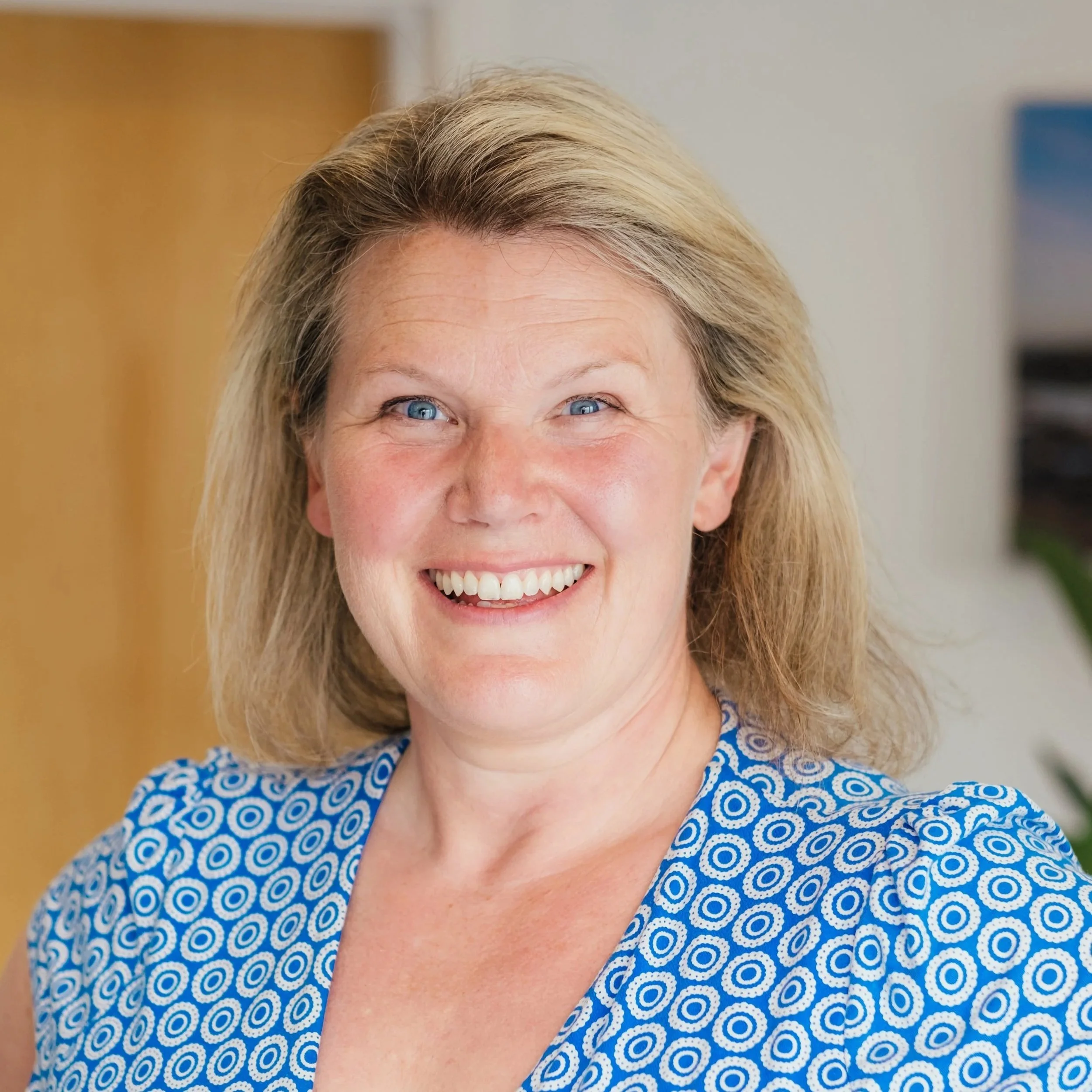 A smiling middle-aged woman with blonde hair and blue eyes, wearing a blue patterned blouse, standing in an indoor setting.