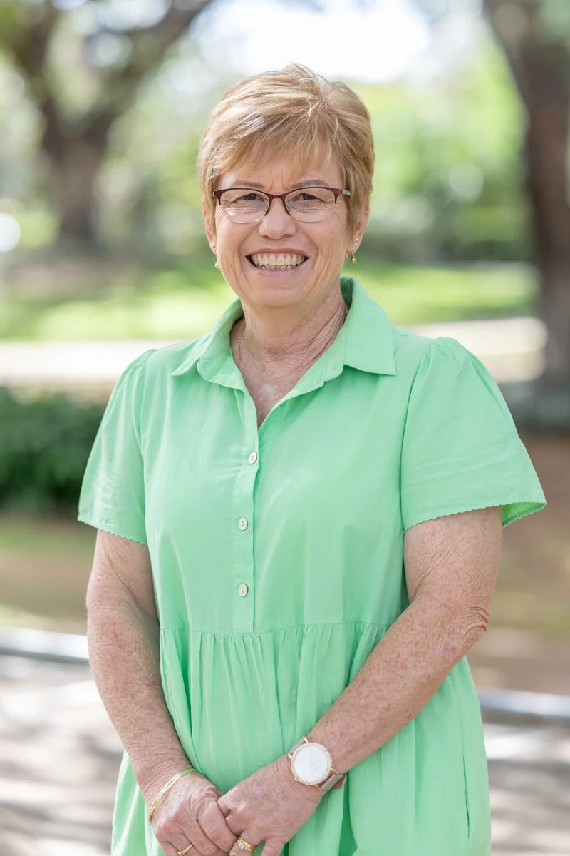 A smiling middle-aged woman with short light brown hair and glasses, wearing a light green dress, standing outdoors with a blurred background of trees and greenery.