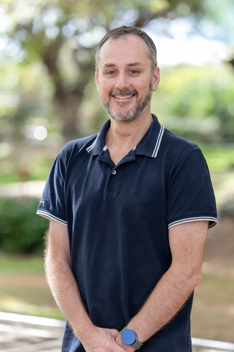 A man with short brown hair and a beard smiling at the camera, wearing a navy blue polo shirt with white stripes on the collar and sleeves, standing outdoors with blurred trees and greenery in the background.