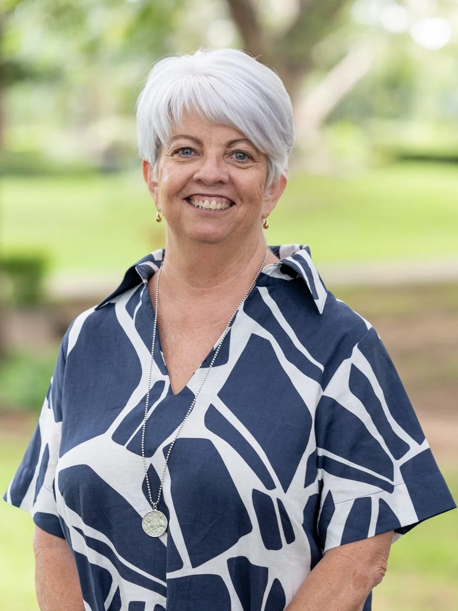 A smiling woman with short white hair wearing a colorful floral and patterned blouse, standing outdoors with a background of trees and sunlight.