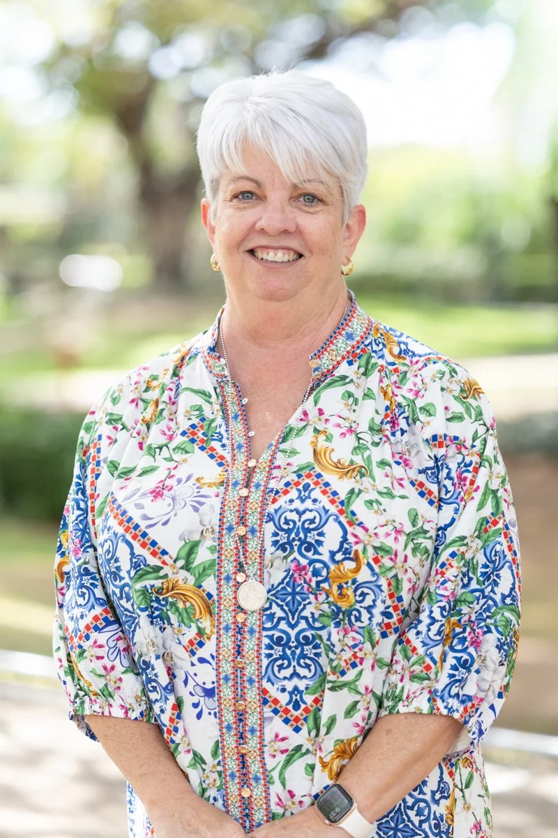 A smiling woman with short white hair wearing a colorful floral and patterned blouse, standing outdoors with a background of trees and sunlight.