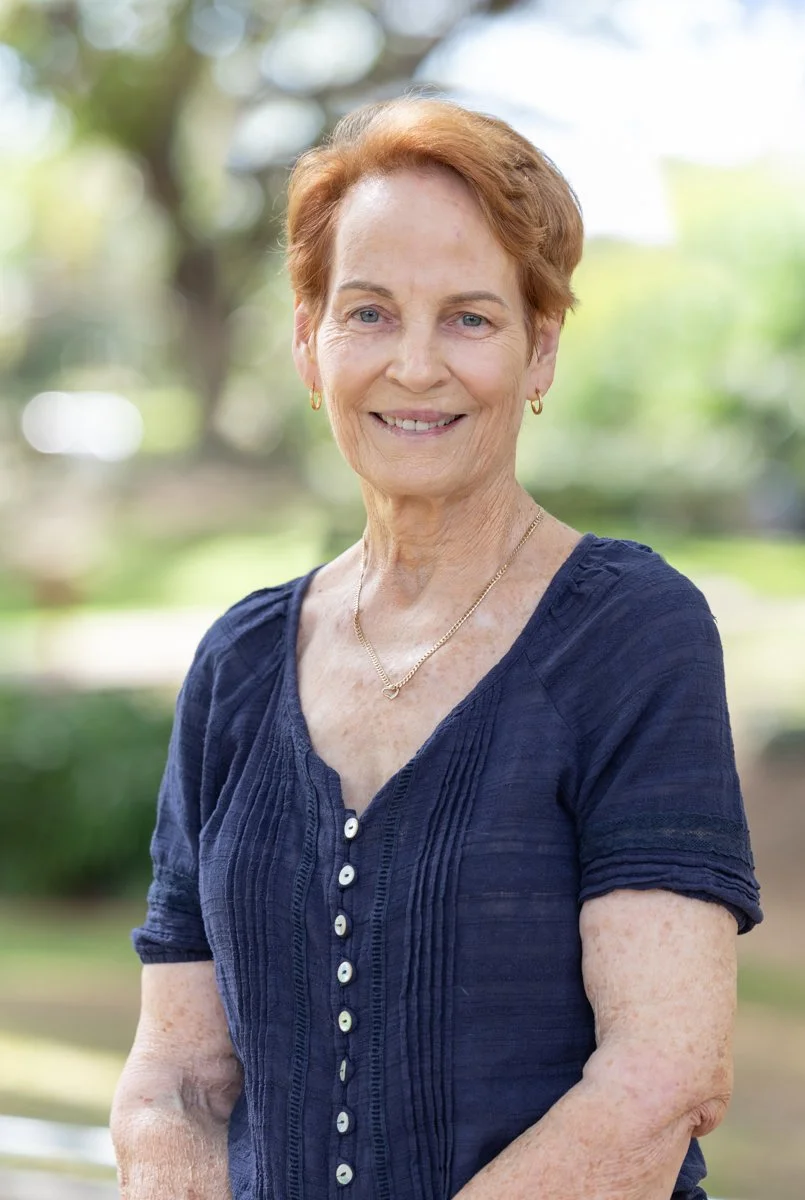 A smiling elderly woman with short red hair outdoors, wearing a navy blue blouse with buttons, gold earrings, and a gold necklace, in a park with blurred trees in the background.