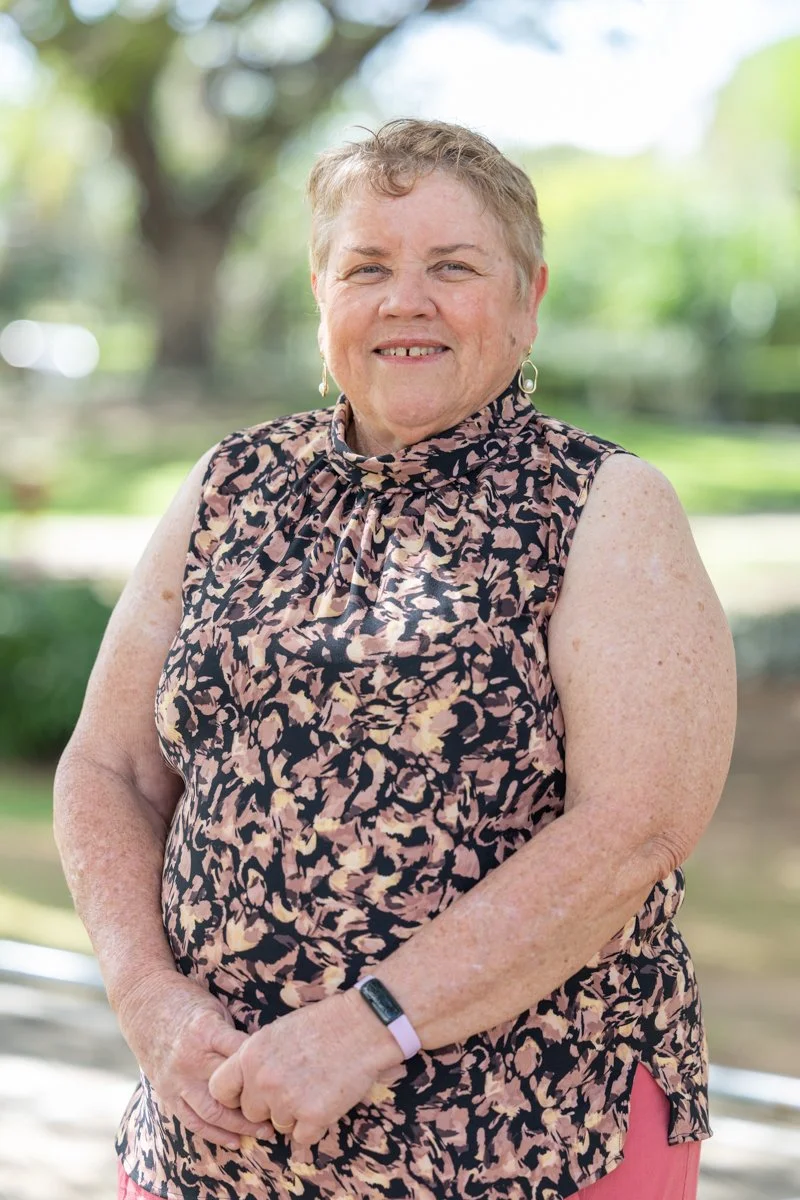 A smiling middle-aged woman with short, curly blonde hair standing outdoors, wearing a sleeveless animal print blouse. The background is blurred greenery and trees.