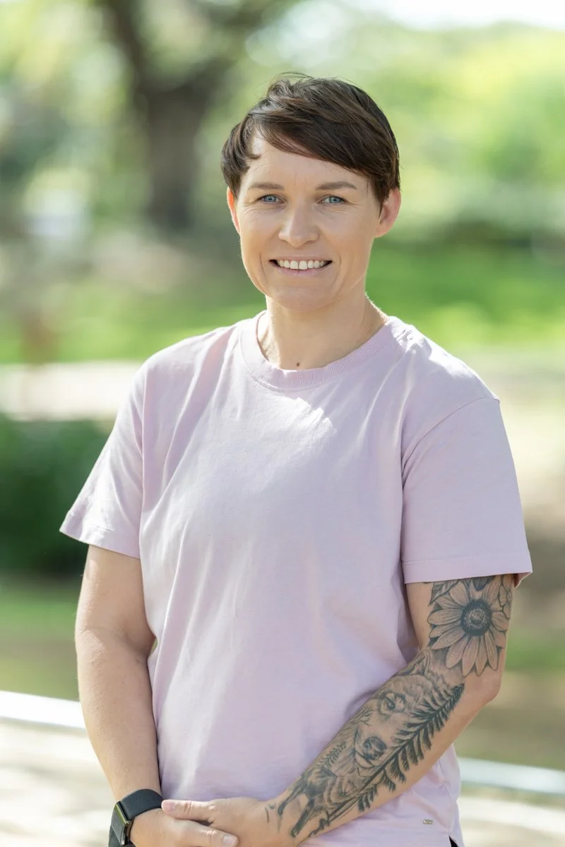 Woman with short dark hair, wearing a light pink T-shirt, smiling, with a tattoo sleeve on her left arm, outdoors with blurred greenery in the background.