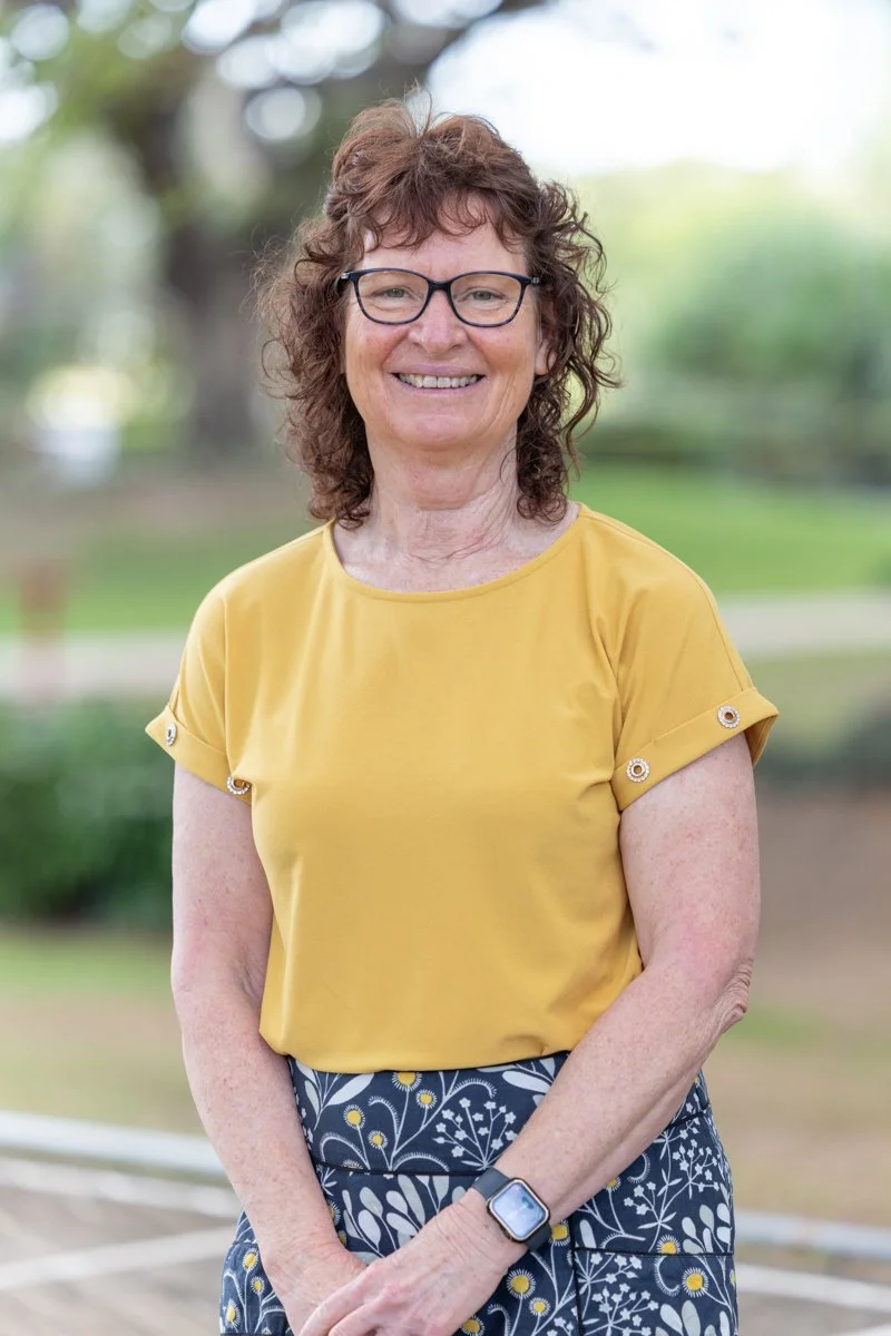 A woman with curly brown hair, glasses, and a cheerful expression, standing outside in a park-like setting with a blurred green background, wearing a yellow top with rolled sleeves and a floral skirt.