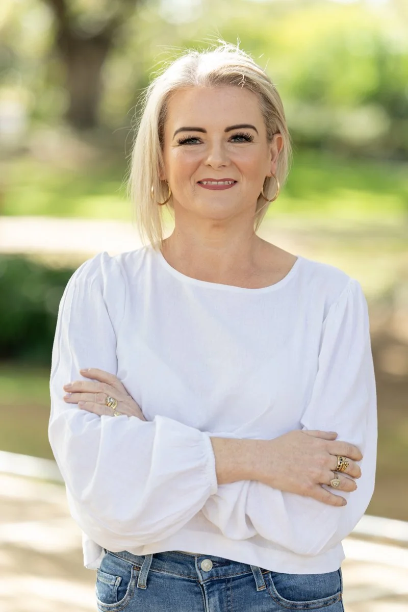 A woman with blonde hair, wearing a white long sleeve blouse and blue jeans, standing outdoors with arms crossed and smiling at the camera.