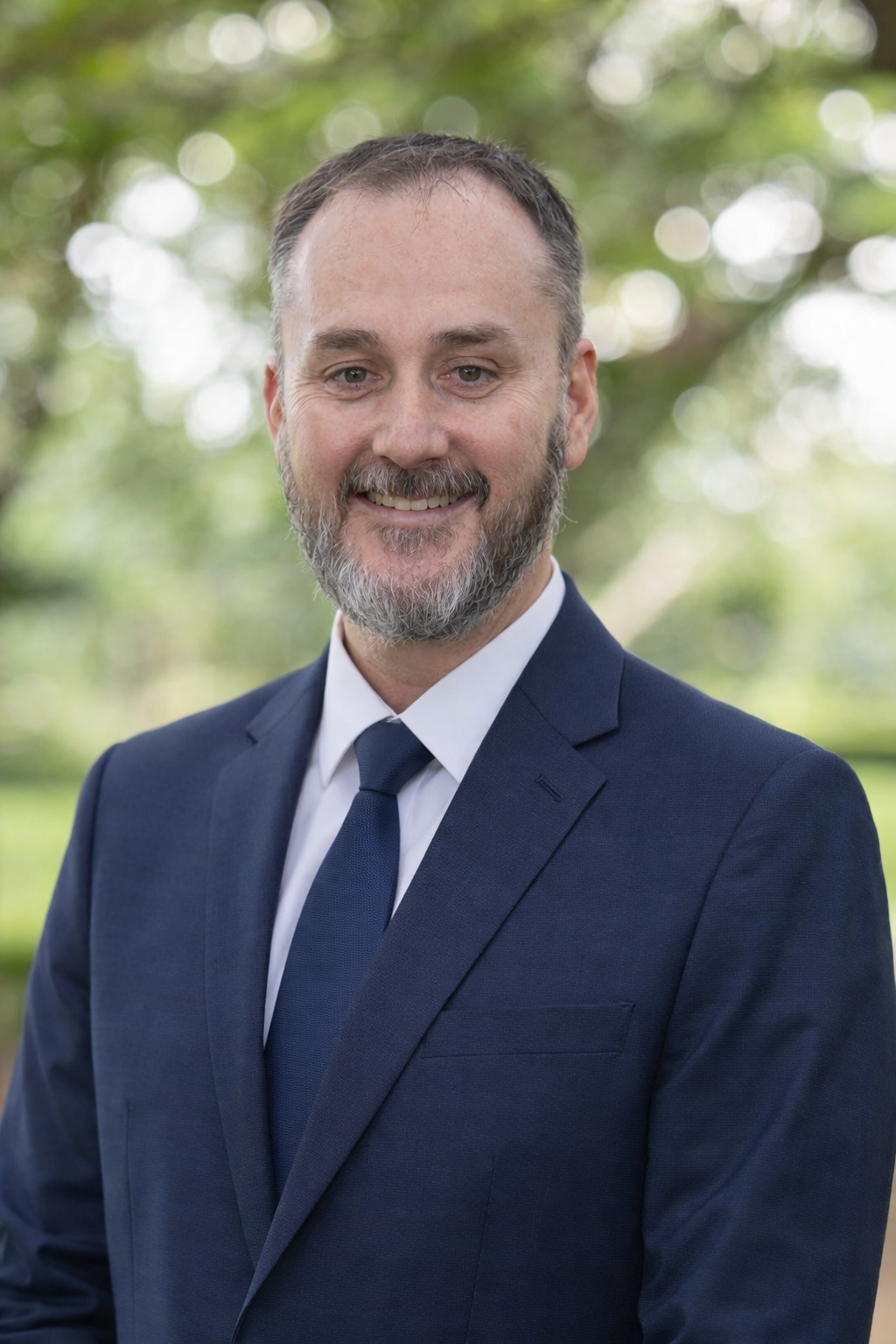 A man with short brown hair and a beard smiling at the camera, wearing a navy blue polo shirt with white stripes on the collar and sleeves, standing outdoors with blurred trees and greenery in the background.