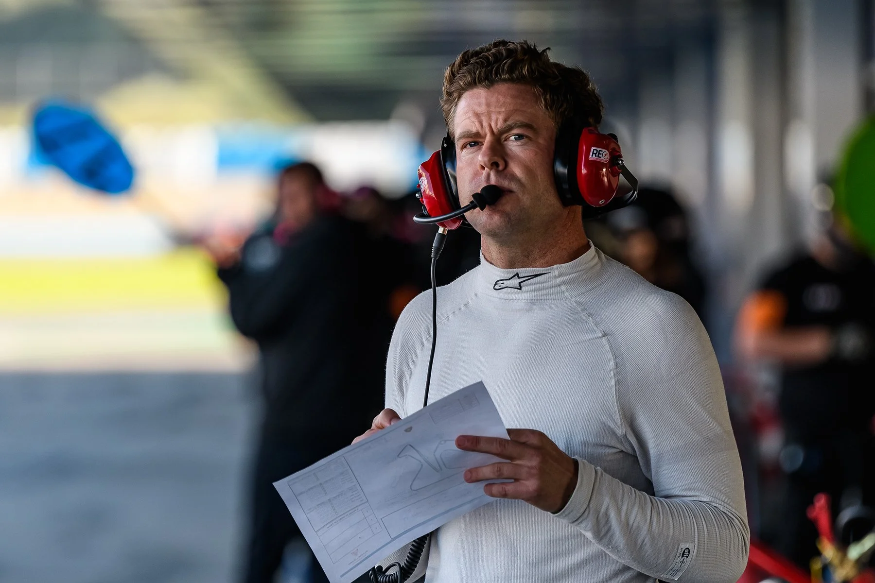 Race car driver standing in pit lane, wearing a white racing suit and a red headset, holding a sheet of paper, with blurred background of team members and garage.