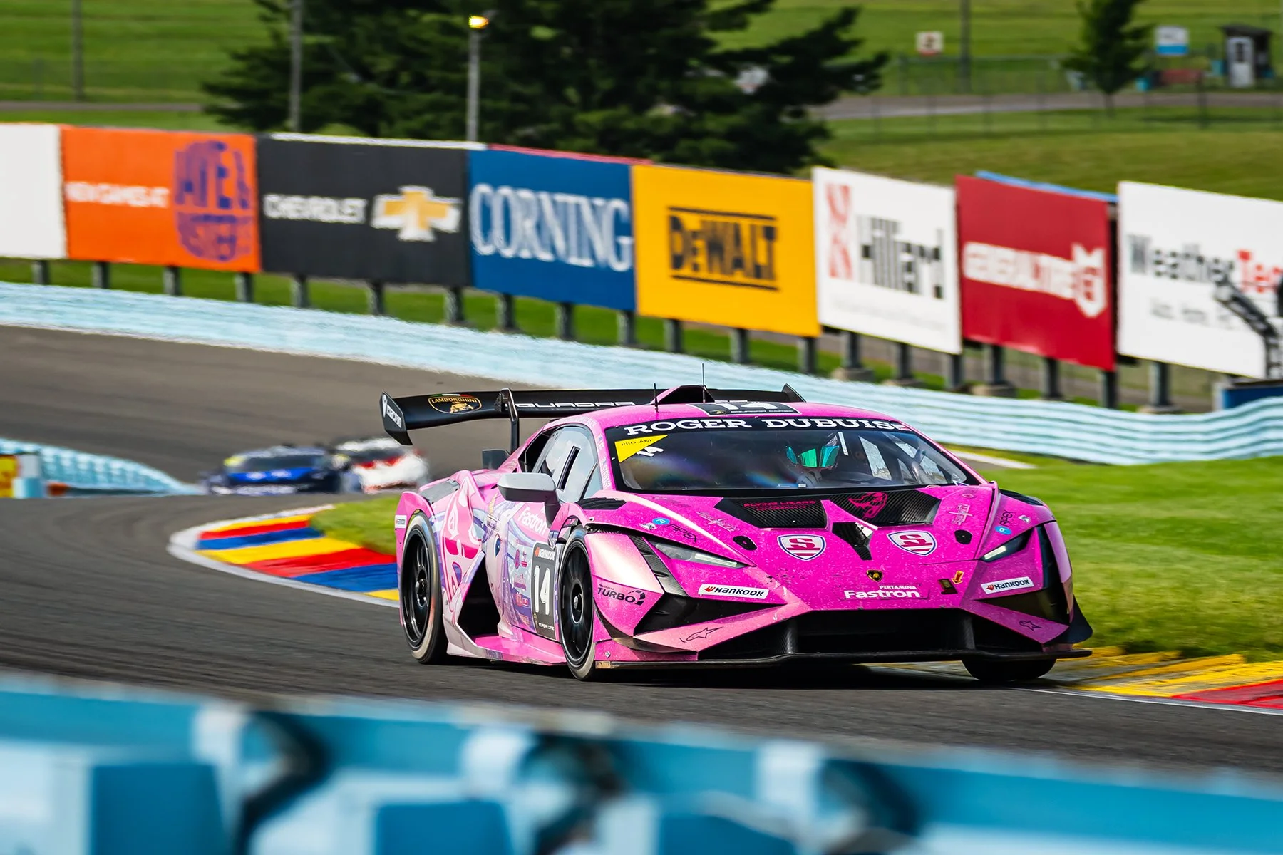 Pink racing car navigating a turn on a race track, with racing advertisements and barriers in the background.