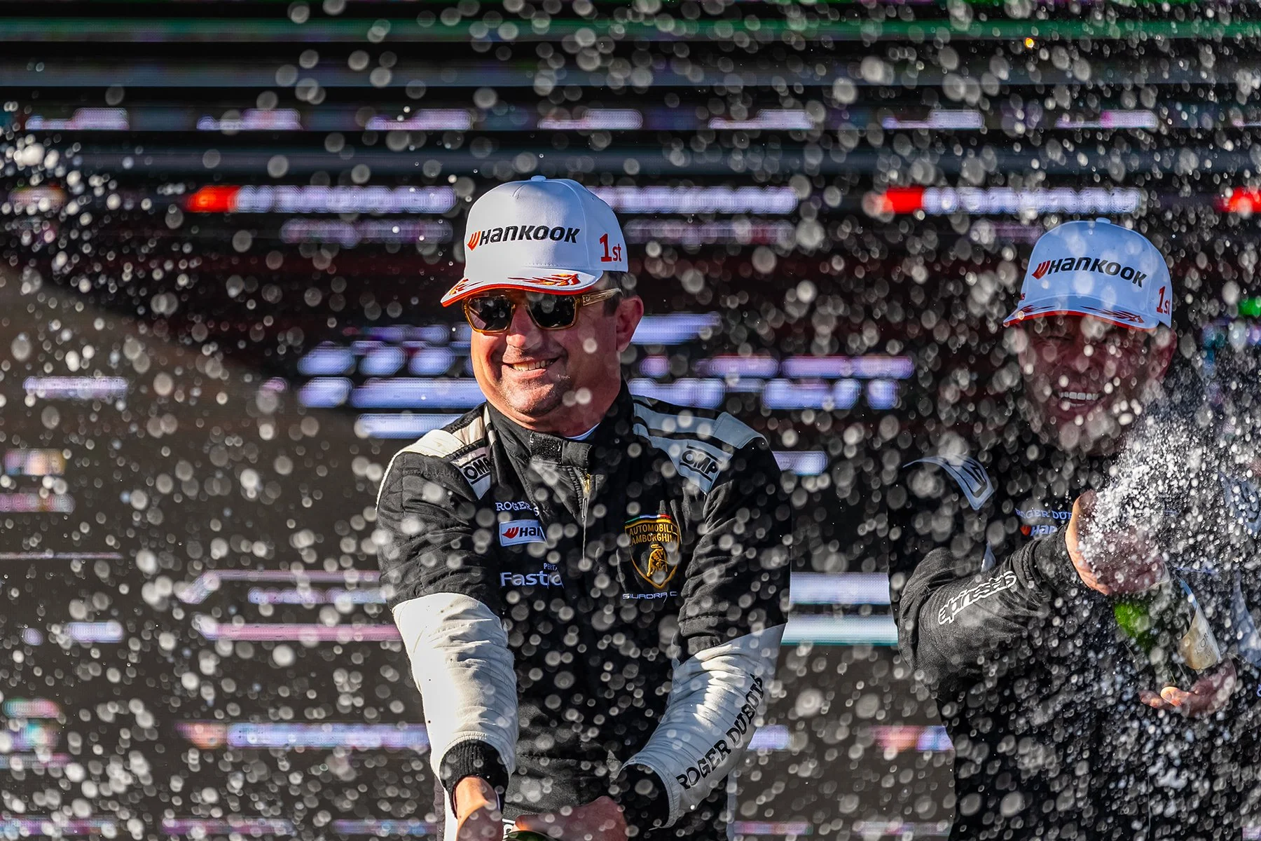 Race car drivers celebrating on the podium, spraying champagne, wearing racing suits and white caps with 'Hankook' logos, standing in front of a backdrop with blurred racing-related graphics.
