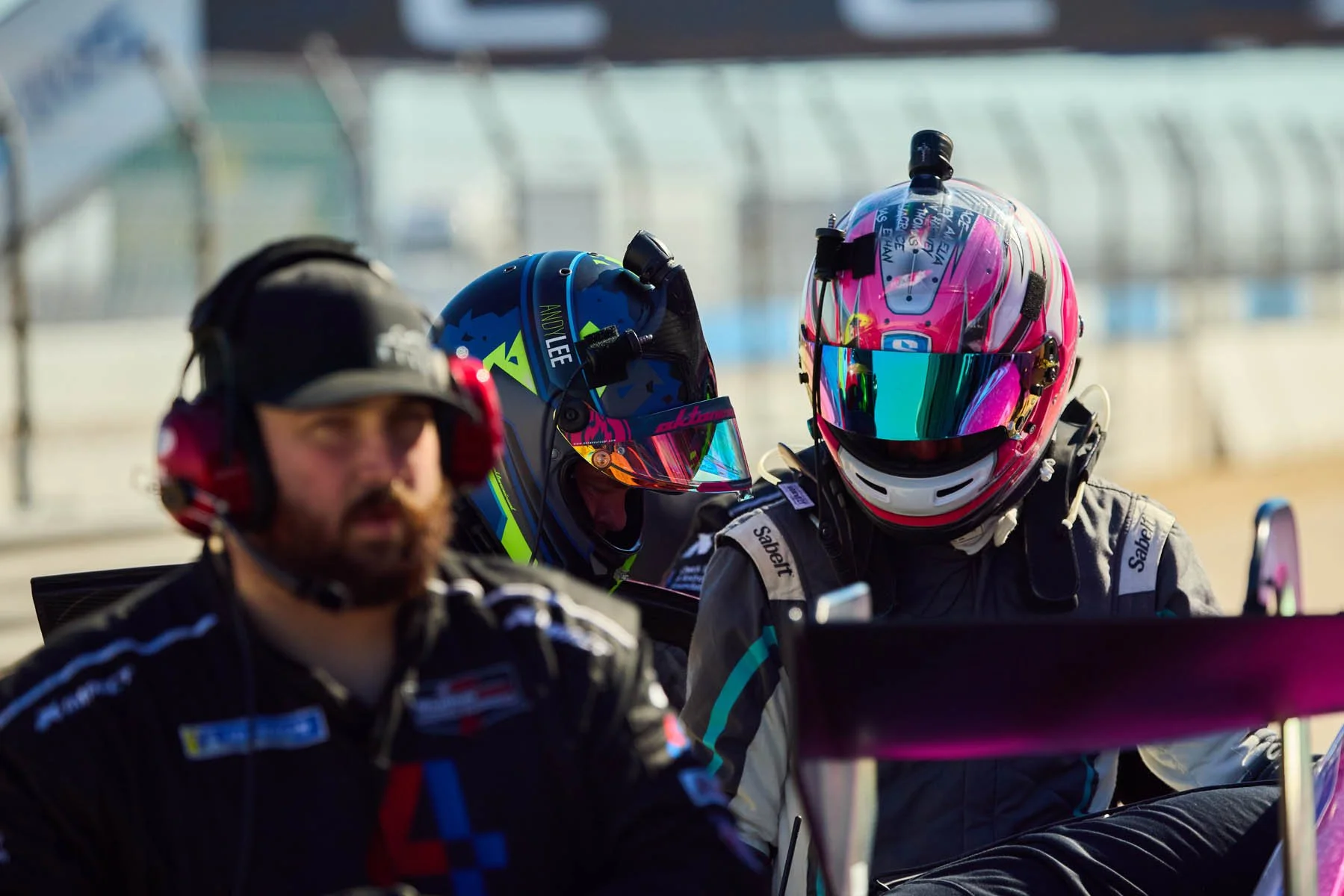 Three individuals, two wearing racing helmets and suits, and one man wearing headphones and a racing suit, are in a racing environment.
