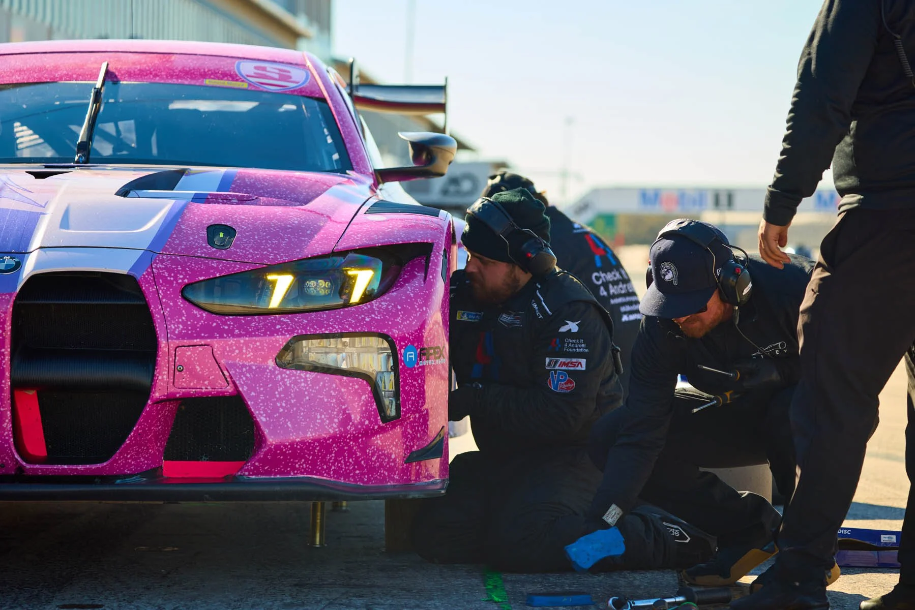 Race car pit crew working on a pink race car during a race, with team members wearing headsets and safety gear.