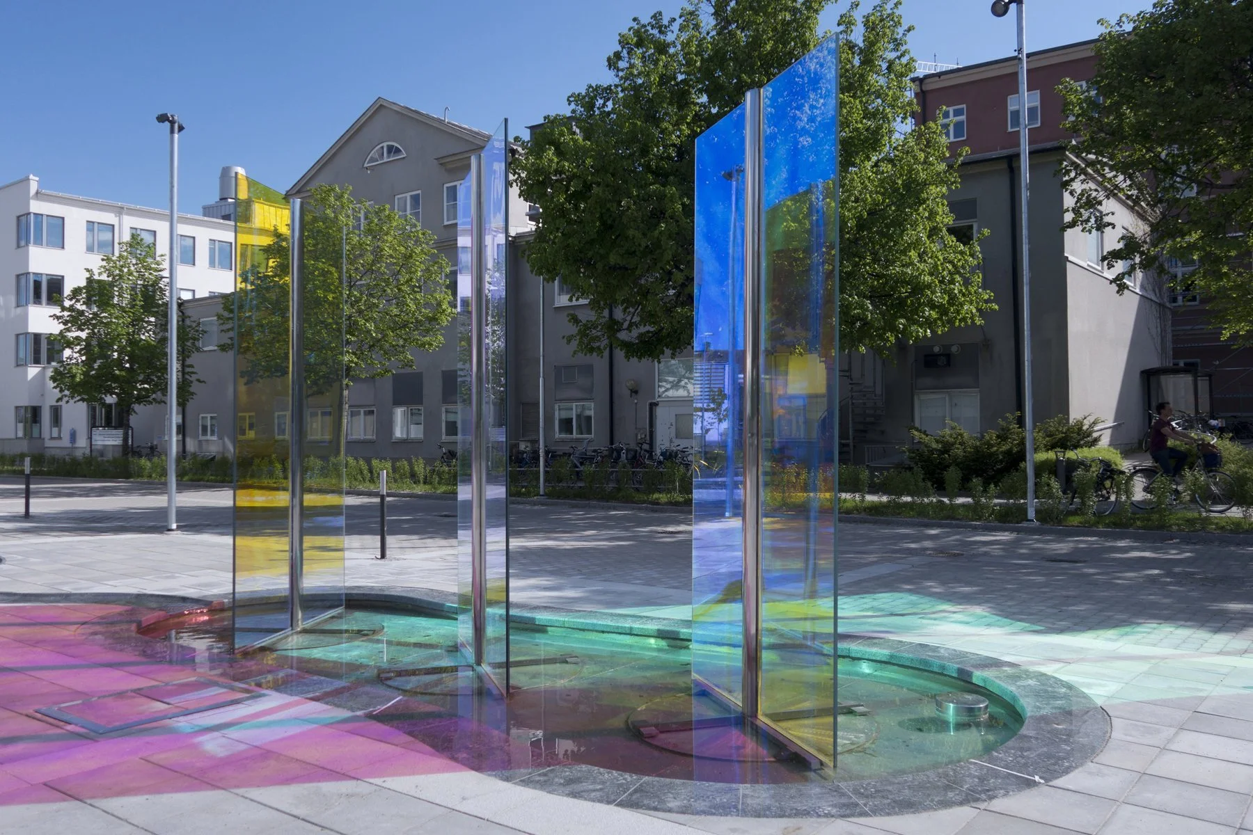 Colorful glass art installation outdoors on a sunny day, with trees, apartment buildings, and a person biking in the background.