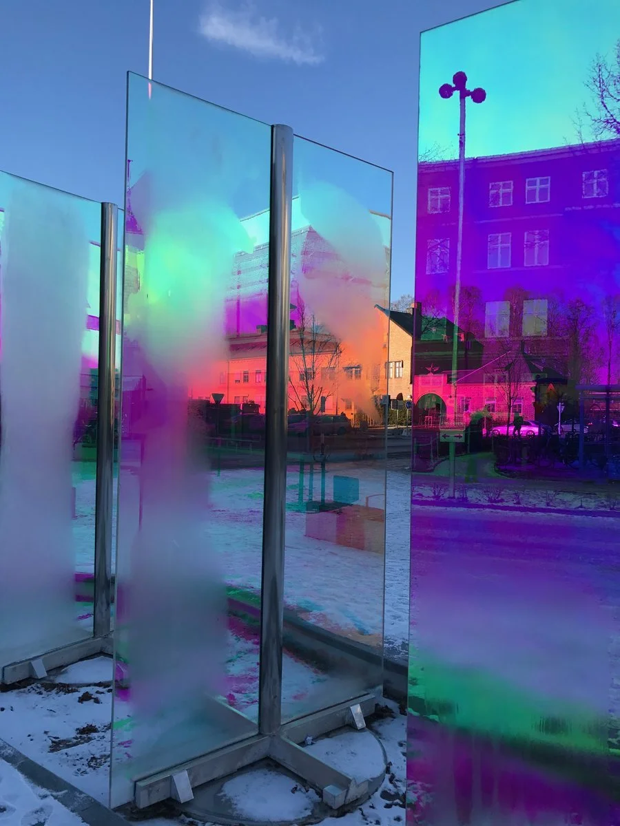 Colorful mirrored art installation outdoors during snowy day, with buildings and trees reflected in the surfaces.