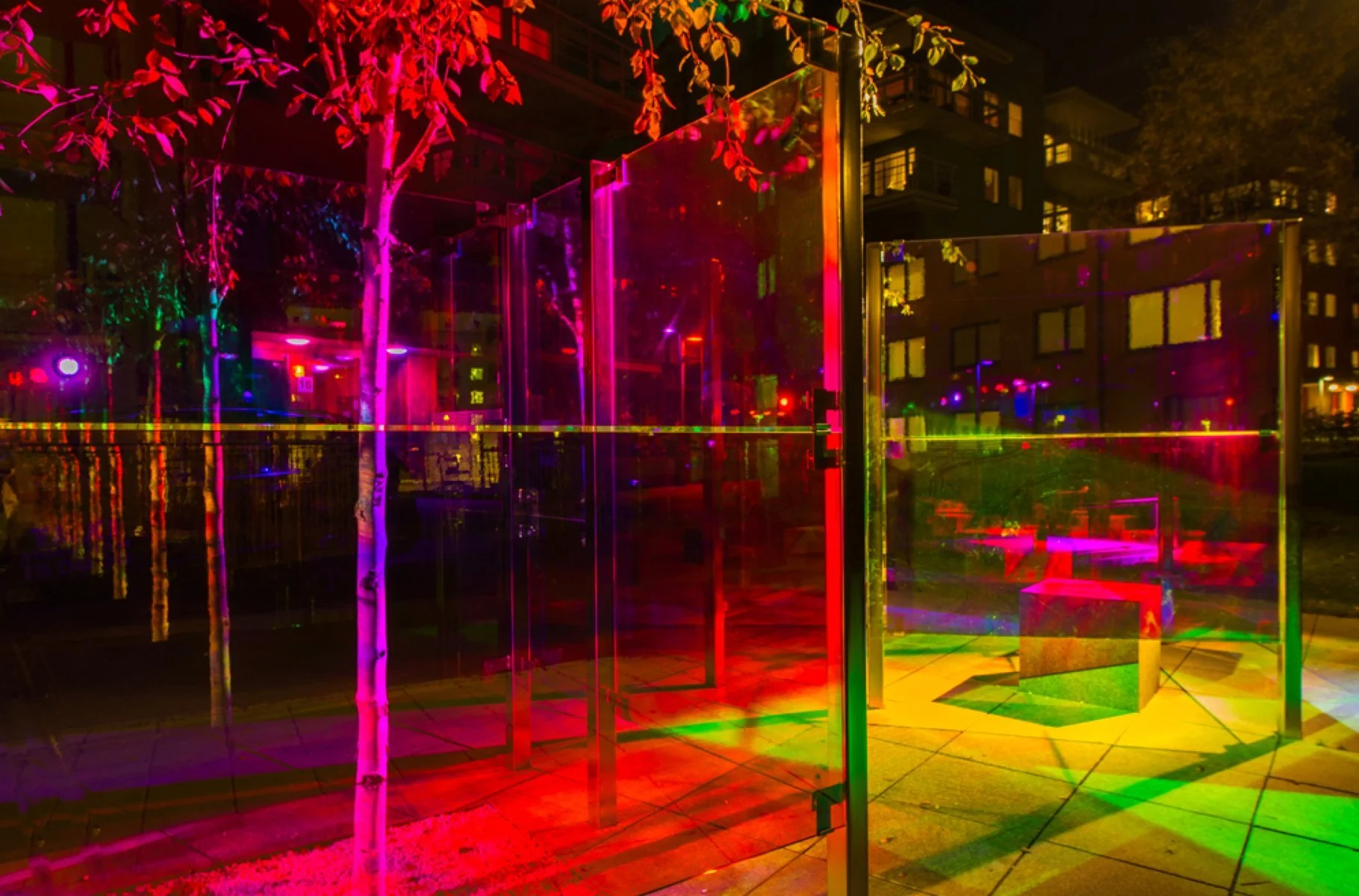 Night scene at an outdoor art installation with colorful neon lights, glass panels, a small tree, and a bench, in an urban area with buildings in the background.