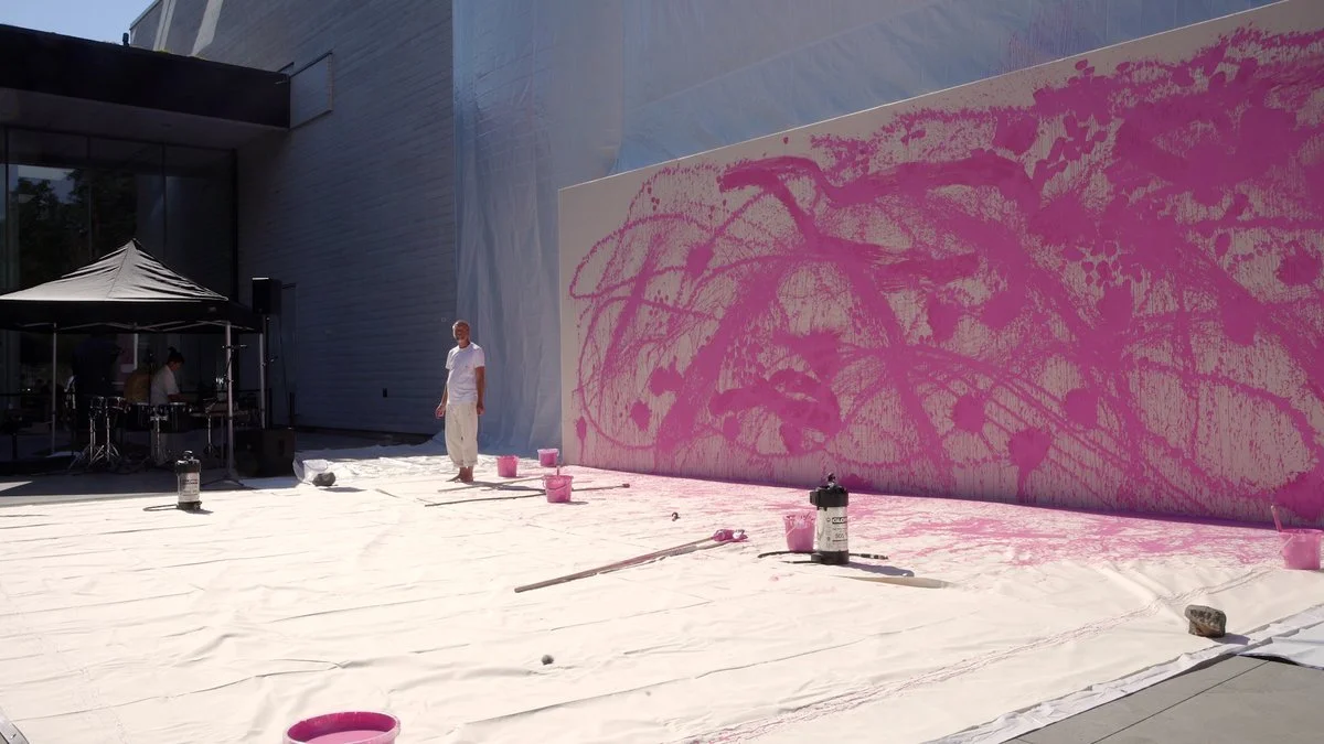 An artist painting a large outdoor canvas with pink paint, standing on a white protective sheet, while people work under a black canopy to the side.