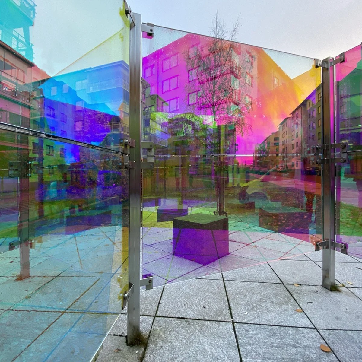 Colorful glass panel fence reflecting nearby trees and buildings in an urban setting with overcast sky.