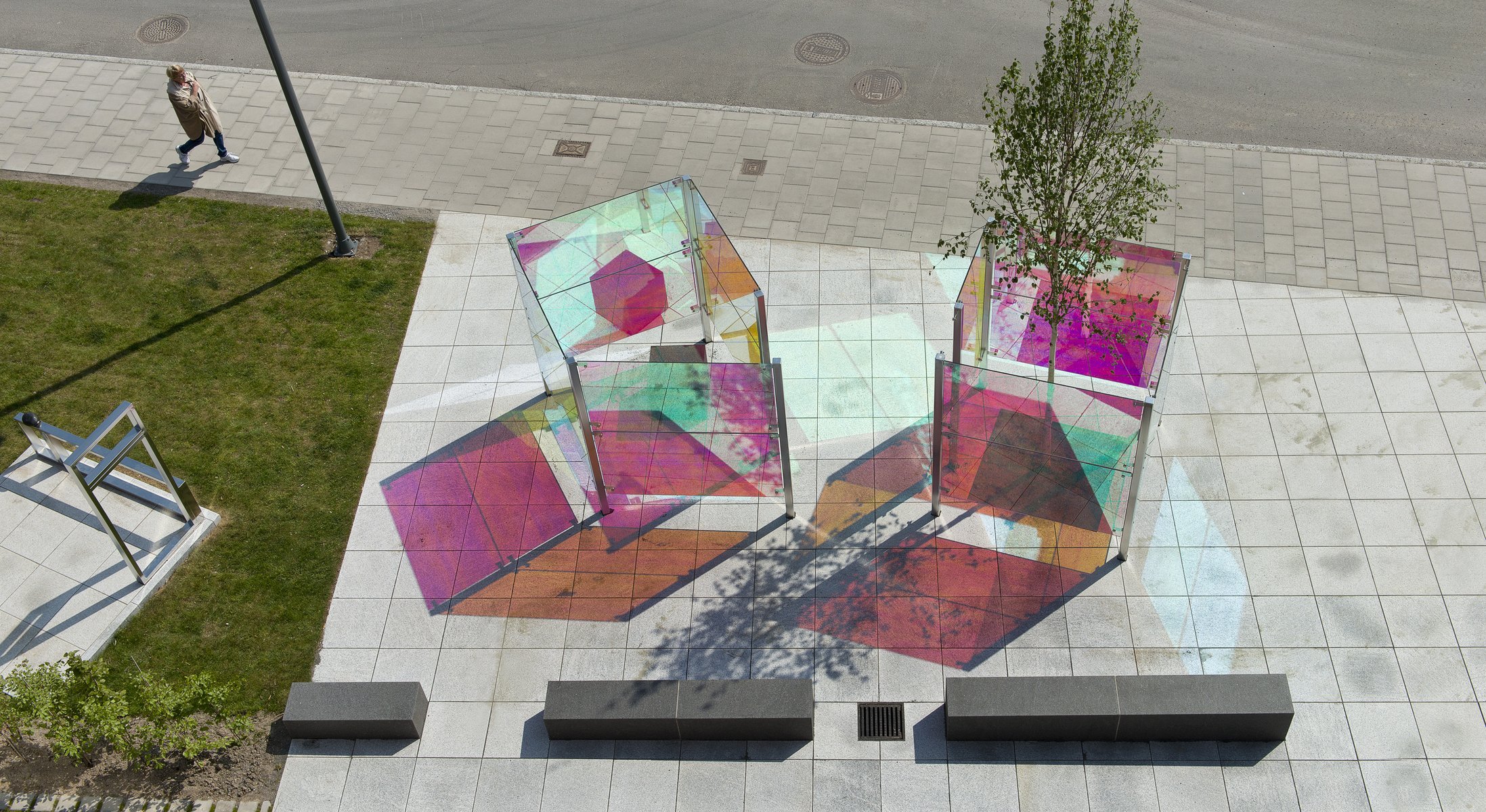 A colorful modern art installation with interconnected glass panels on a city sidewalk, casting vibrant shadows and reflections. A person walks by on the street, and a small tree and green grass are nearby.