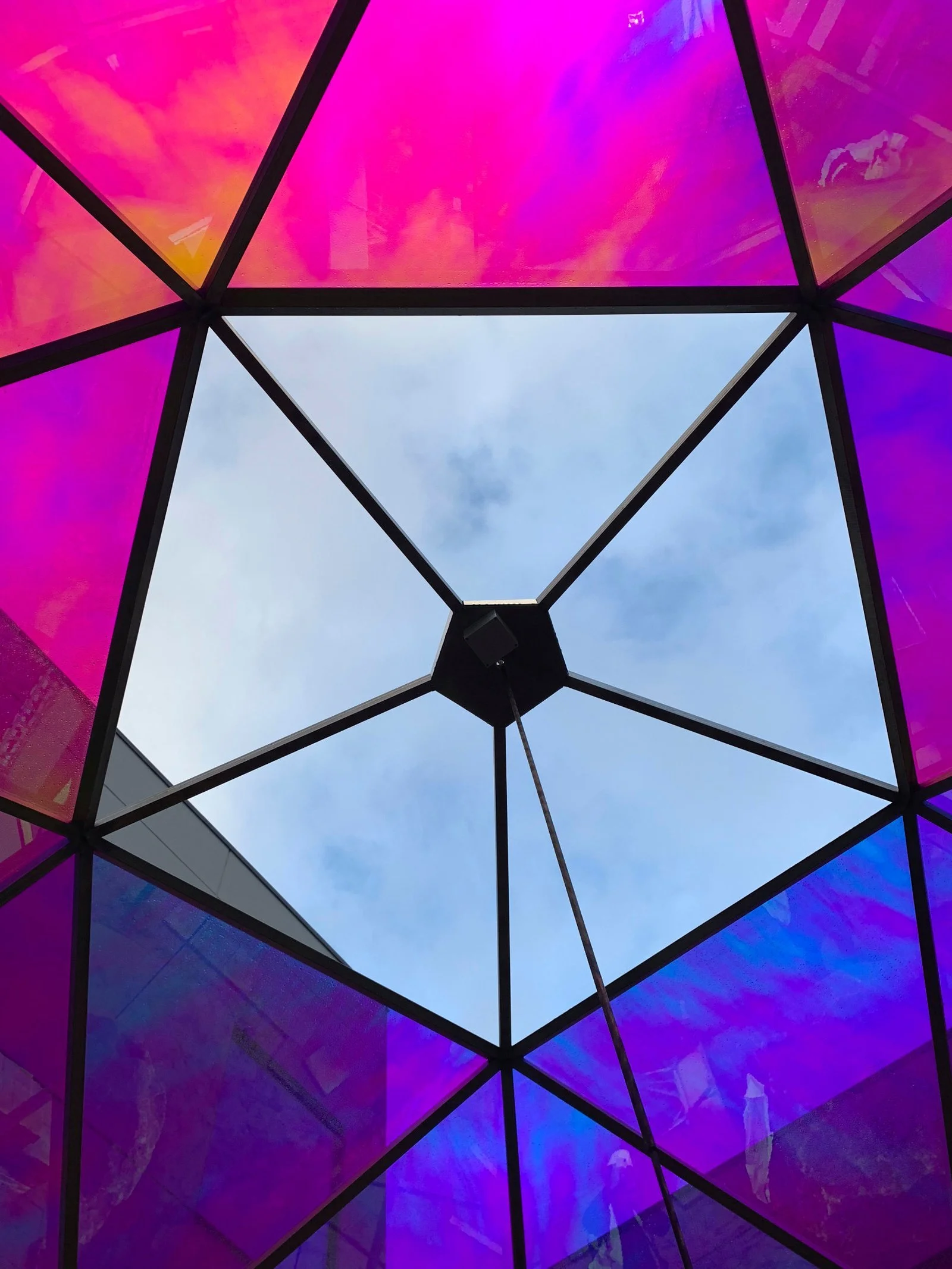 Looking up at a geometric glass ceiling with colorful pink, purple, and blue panels and a central square opening showing a cloudy sky.