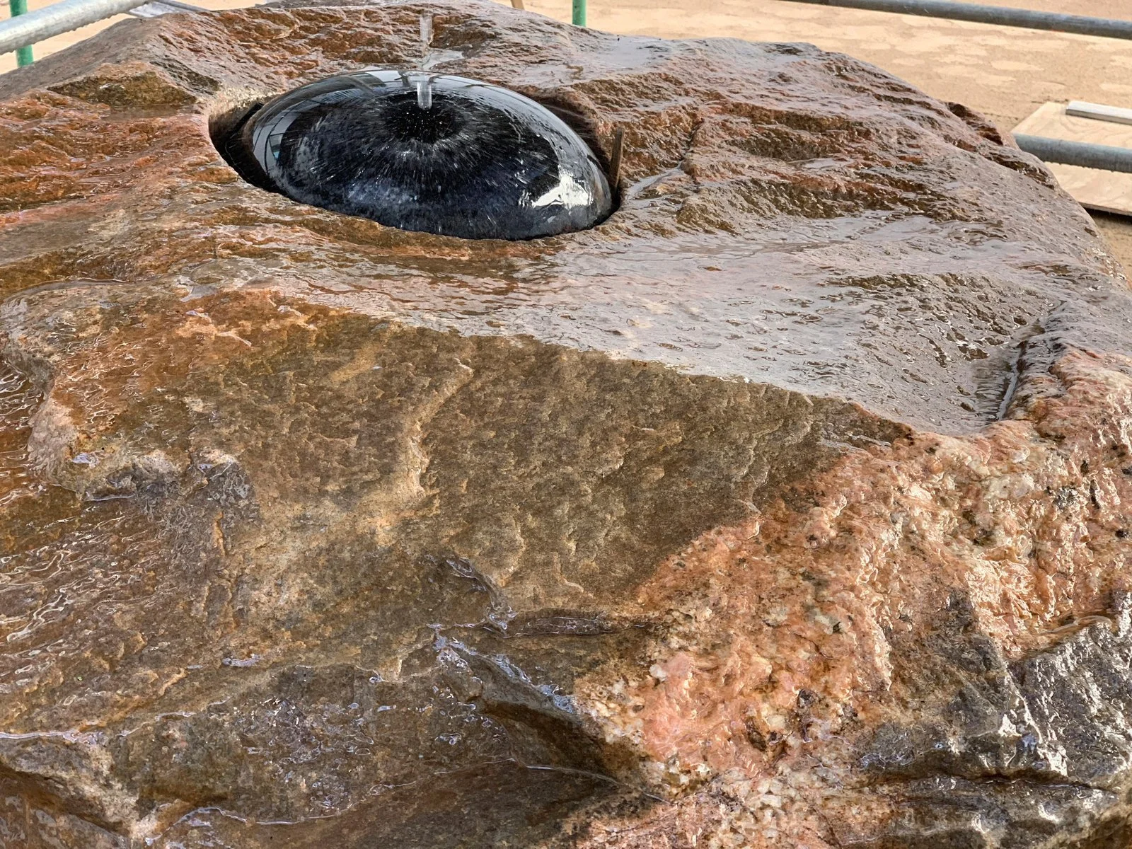 Close-up of a water fountain feature with a black, dome-shaped element from which water is spraying upward, set on a rough, natural-looking stone surface.
