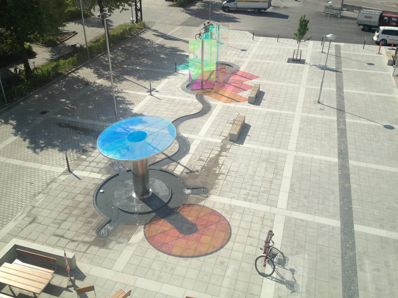 Empty outdoor plaza with colorful rainbow-shaped glass sculptures, benches, and a bicycle parked on the paved surface.