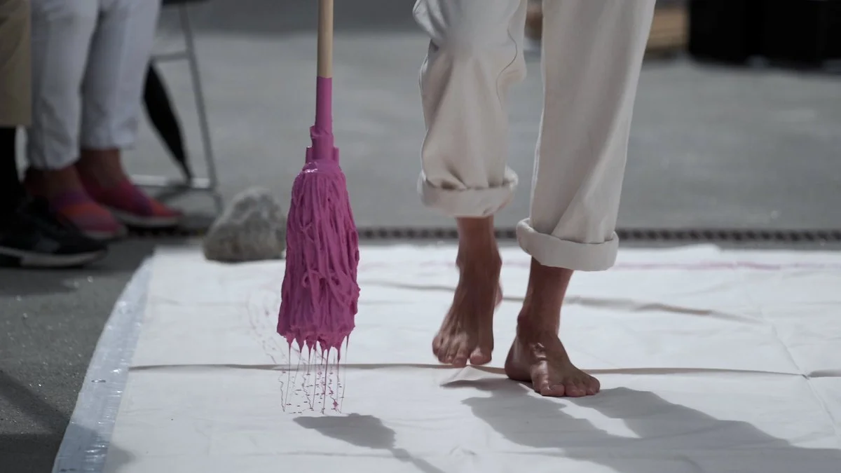 Person walking barefoot on white cloth, holding a pink mop with pink paint dripping from it.