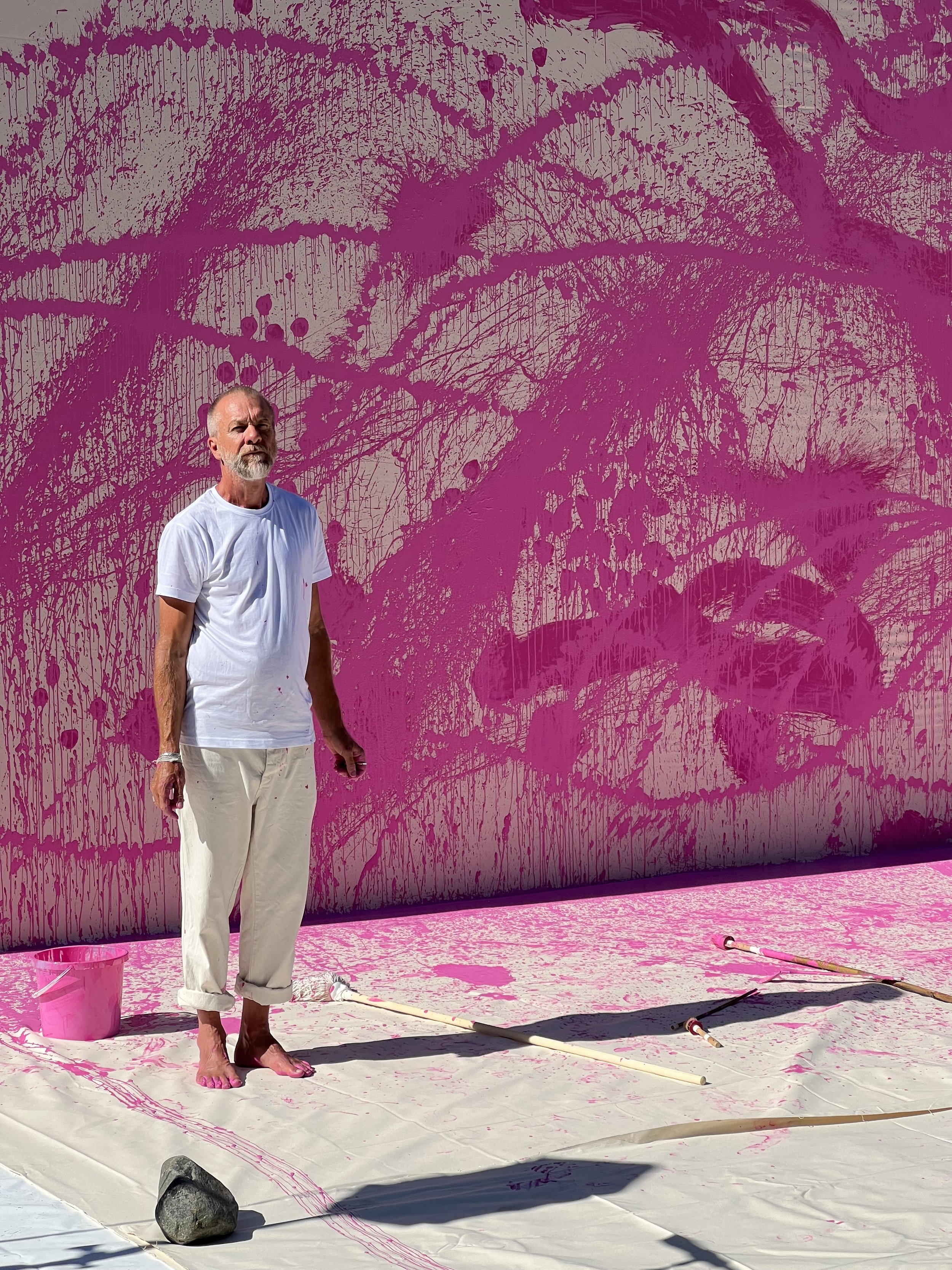 The artist Torbjörn Johansson wearing a white t-shirt and beige pants stands on a white surface covered in pink paint. Behind him is a large wall, painted with paint drips and splatters, A pink bucket, and paint tools are on the ground.