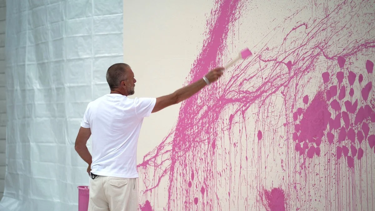 A man wearing a white t-shirt and light-colored pants is painting a wall with pink paint, creating an abstract pattern with splashes and streaks.