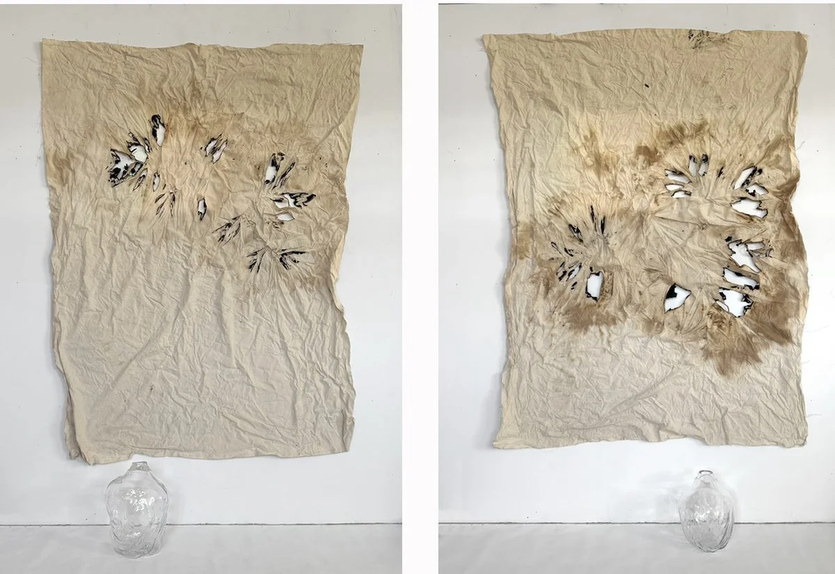 Two crumpled, burnt pieces of paper mounted on a wall, each with multiple holes, with a clear glass jar placed below them.