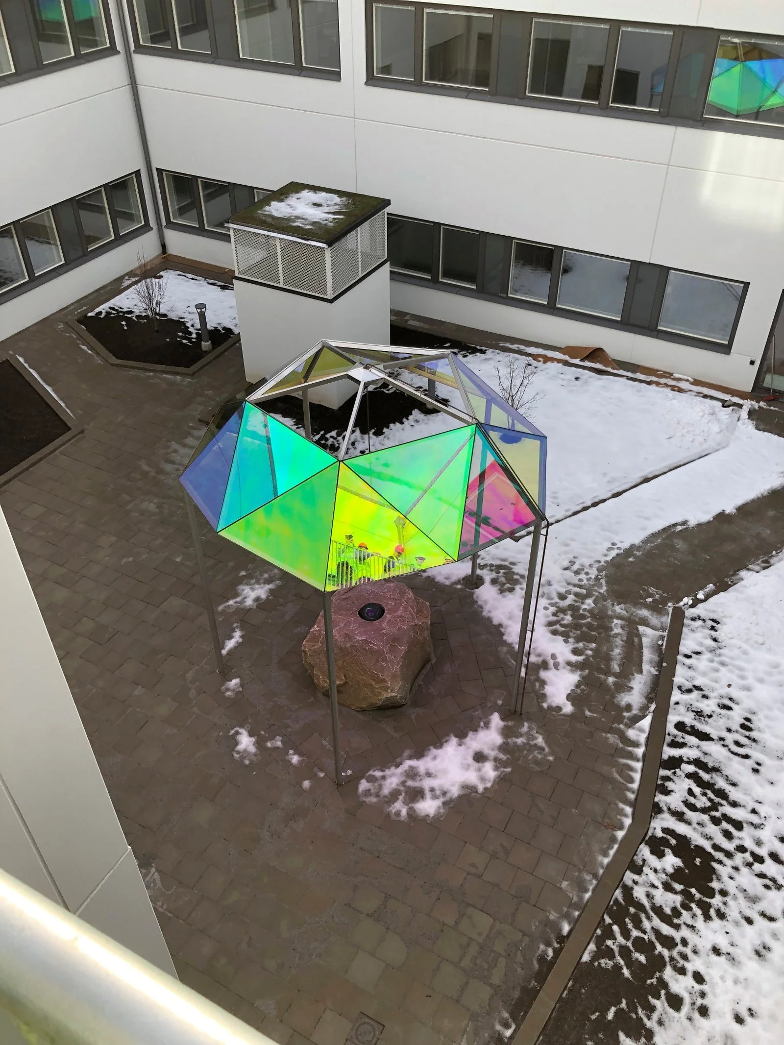 A rooftop courtyard with snow patches, a large rock, and a modern, colorful glass geometric pavilion covering a small well or fountain.
