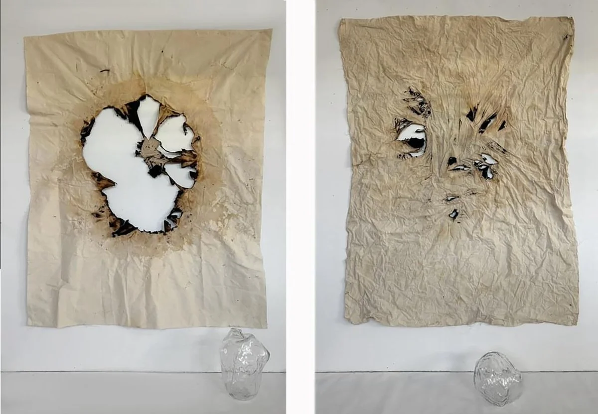 Two pieces of crumpled paper with burn marks and holes, mounted on a wall, with a clear plastic container below each piece.